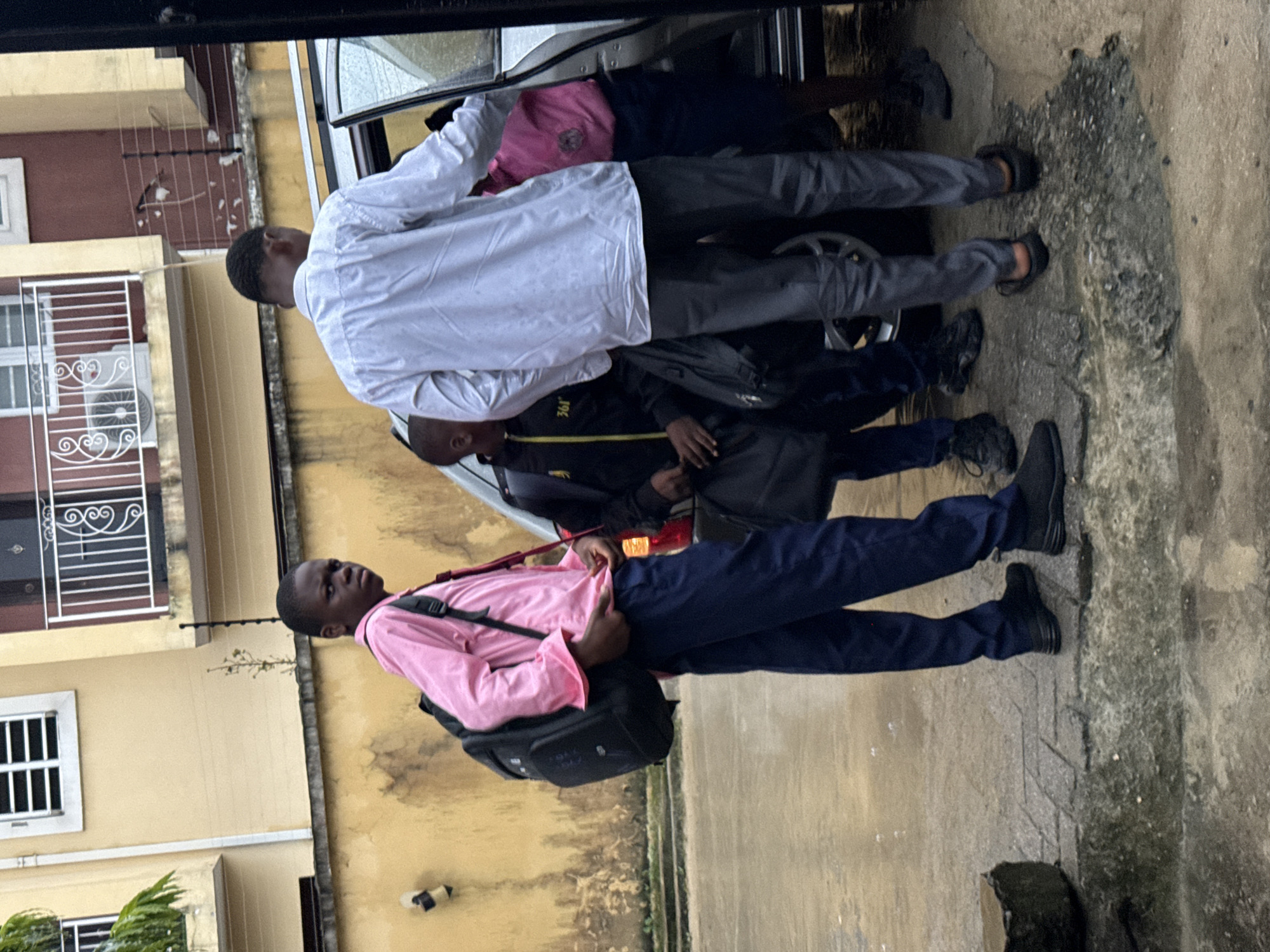 Four individuals, three young males and one possibly older male, are gathered near the open door of a silver sedan. The young males are wearing school uniforms, with two in blue trousers and pink or yellow shirts, and one in black trousers and a yellow and black jacket. One of the young males in blue trousers and a pink shirt is looking directly at the camera. The older male, wearing a white shirt and gray trousers, appears to be assisting the others into the vehicle. They are carrying backpacks. The ground is wet and appears to be paved with bricks or stones, suggesting an outdoor setting. The background shows a building with yellow and reddish-brown walls, and a balcony with a white railing. The overall impression is that of students getting into a car, possibly after school, in a residential or urban environment in Ado, Nigeria, as indicated by the presence of school uniforms and a typical Nigerian building facade. The presence of water on the ground suggests it may have recently rained.