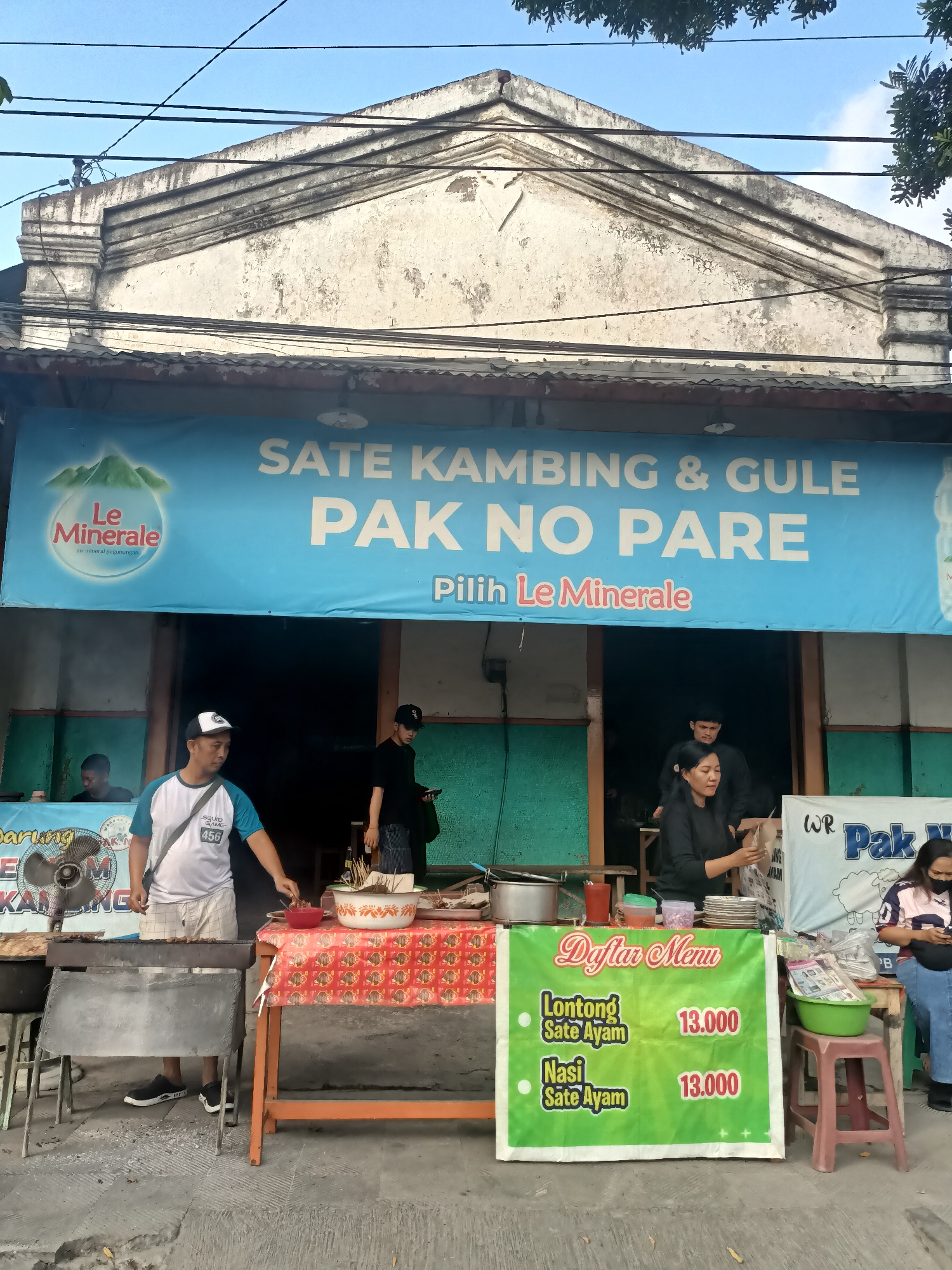 This is an eye-level medium shot of a street food stall in Pare, Indonesia. The stall, named "Sate Kambing & Gule Pak No Pare," displays a large blue banner with white text advertising their offerings. To the left of the banner is the Le Minerale logo, a brand of bottled water. The stall is set up on a paved area in front of a building with a white, weathered facade and a corrugated metal roof.
Several people are visible. A man wearing a blue and white t-shirt and plaid shorts stands by a grill, holding tongs and appearing to cook. Another man in a black t-shirt and cap stands to the right, holding a phone. A woman in a black top is actively serving or preparing food behind a table covered with a red tablecloth. To the far right, a woman in a patterned shirt and blue jeans is standing, looking down.
In the foreground, a green menu board lists "Lontong Sate Ayam" and "Nasi Sate Ayam," both priced at 13,000. Various food preparation items, including bowls of food, skewers, plates, and cooking utensils, are arranged on tables. The overall environment suggests a busy street-side food establishment.