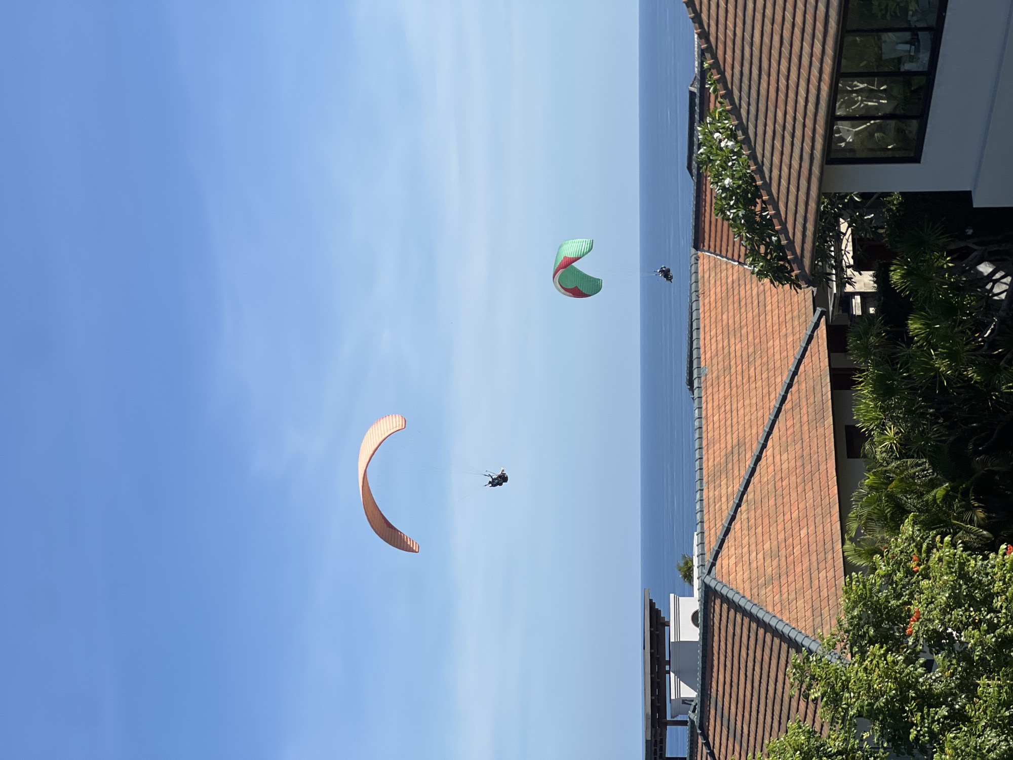 The image shows a view of the sky above Unknown, Unknown, with paragliders in the air. The sky is blue with some clouds. Two paragliders are visible, one with a peach-colored canopy and the other with a green and red canopy. A coastline is seen in the background. Buildings with orange-tiled roofs are visible on the right side of the frame.