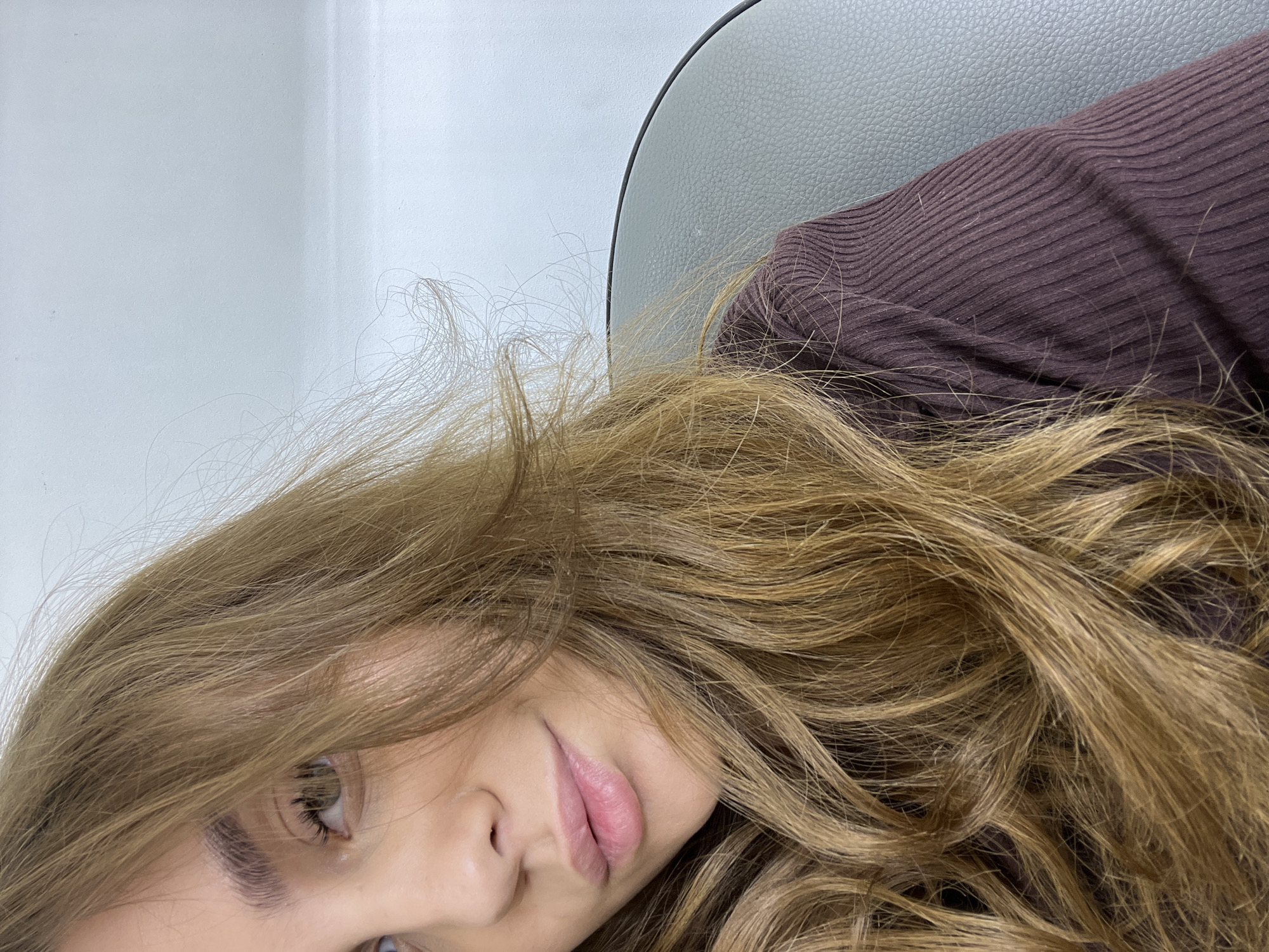 The image shows a young woman's face in close-up, with the camera positioned at an angle. She has light brown hair that is slightly tousled, fair skin, and visible eyelashes and eyebrows. Her lips are closed and appear to be a natural pink color. She is wearing a brown ribbed sweater. In the background, there is a light gray surface, possibly a wall or part of a chair. There is no information about a specific location in Chaikovsky, Russia in the image.