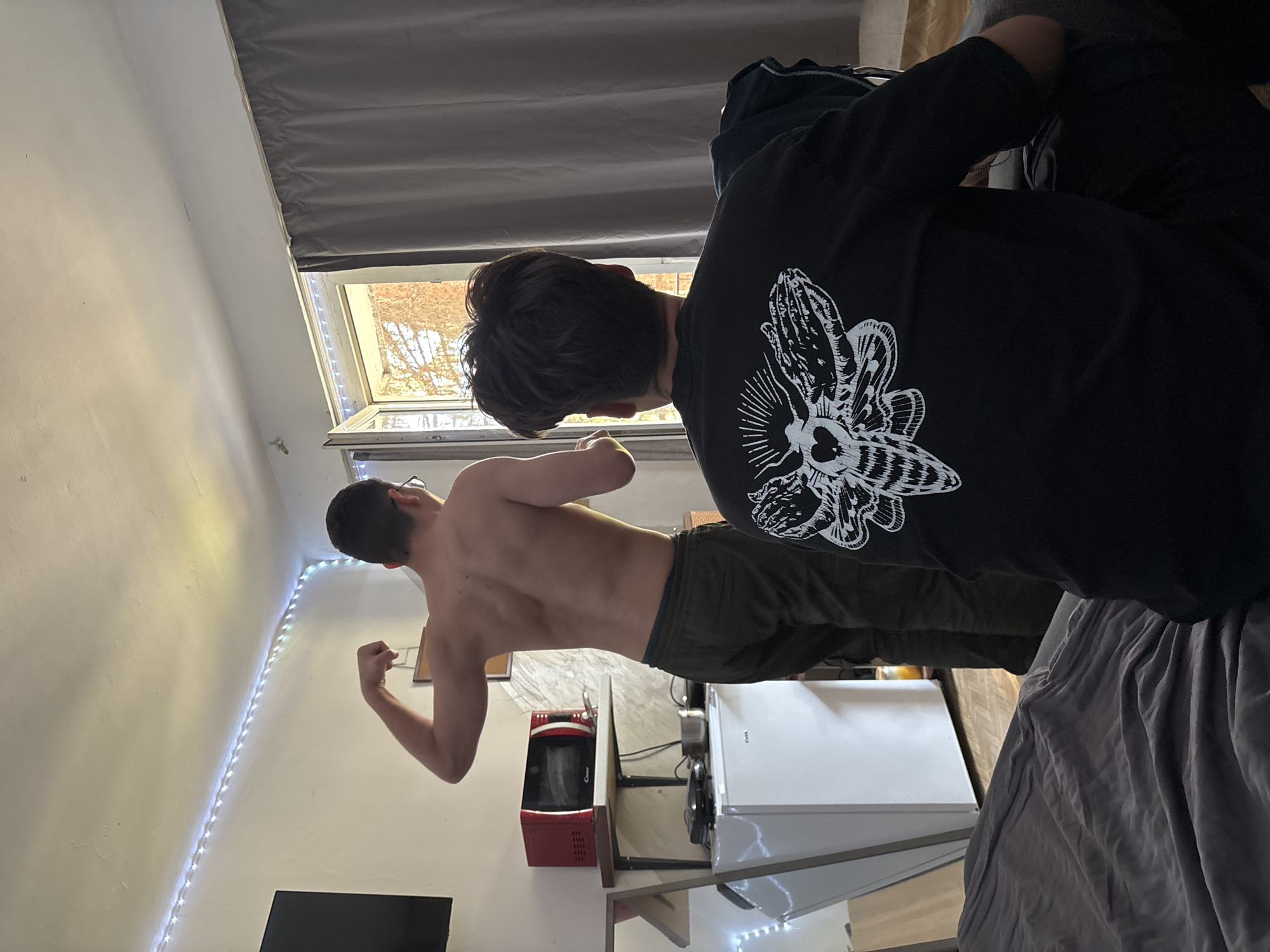 This image depicts two young males in an indoor setting, likely a small room or studio apartment in Troyan, Bulgaria. The shot is taken at a slight angle, with natural light streaming in from a window.
In the foreground, on the right, a male is seen from behind, wearing a black long-sleeved shirt with a prominent white graphic design of a moth or butterfly with a heart-shaped body. He is positioned near a bed or couch covered with rumpled grey fabric.
Further into the background, to the left, another male stands shirtless, also seen from behind, flexing his right bicep. He wears dark-colored pants. His pose suggests a moment of showing off or confidence. The room features plain white walls. A string of bright LED lights is draped along the corner where the wall meets the ceiling, adding a decorative element.
A window in the background reveals an outdoor view of trees, possibly deciduous and bare, suggesting either late autumn or winter. Dark grey blinds or curtains are visible above the window. Below the window, household items such as a red microwave and a white mini-refrigerator are visible, hinting at a compact living arrangement. The overall impression is a casual, everyday scene. No explicit text is visible apart from the graphic on the shirt.