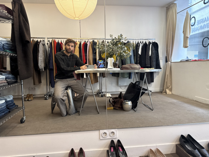 The image depicts the interior of a clothing boutique in Paris, France, as indicated by the reverse reflection of "MAIRIE GYMNASE" visible through a window on the right.
A male individual with a beard and dark hair, dressed in a dark zipped sweater and grey trousers, is seated on a metallic stool. He holds a smartphone horizontally with both hands, looking directly at his reflection in a large mirror that forms the lower part of the wall.
In the midground, a metallic table with sawhorse-style legs holds a white pitcher, a small brown leather bag, a decorative plant arrangement with green leaves and yellow flowers, and a second smartphone displaying a colorful screen. Below the table, a dark tote bag, a brown bag, and a silver-colored pot rest on the brown carpeted floor.
The background features multiple clothing racks displaying various garments. To the far left, a multi-tiered metal shelving unit is stacked with folded denim items, with a dark brown coat hanging from its side. A pair of light brown boots is visible on the floor near this unit. A large, circular, pleated paper lantern hangs illuminated from the ceiling. On the right, a window, partially obscured by a light beige curtain, shows an additional garment hanging on a rod and the reflected external signage. In the foreground, below the mirrored wall, several pairs of women's shoes in black, pink, and beige are arranged on the floor next to white double power outlets.