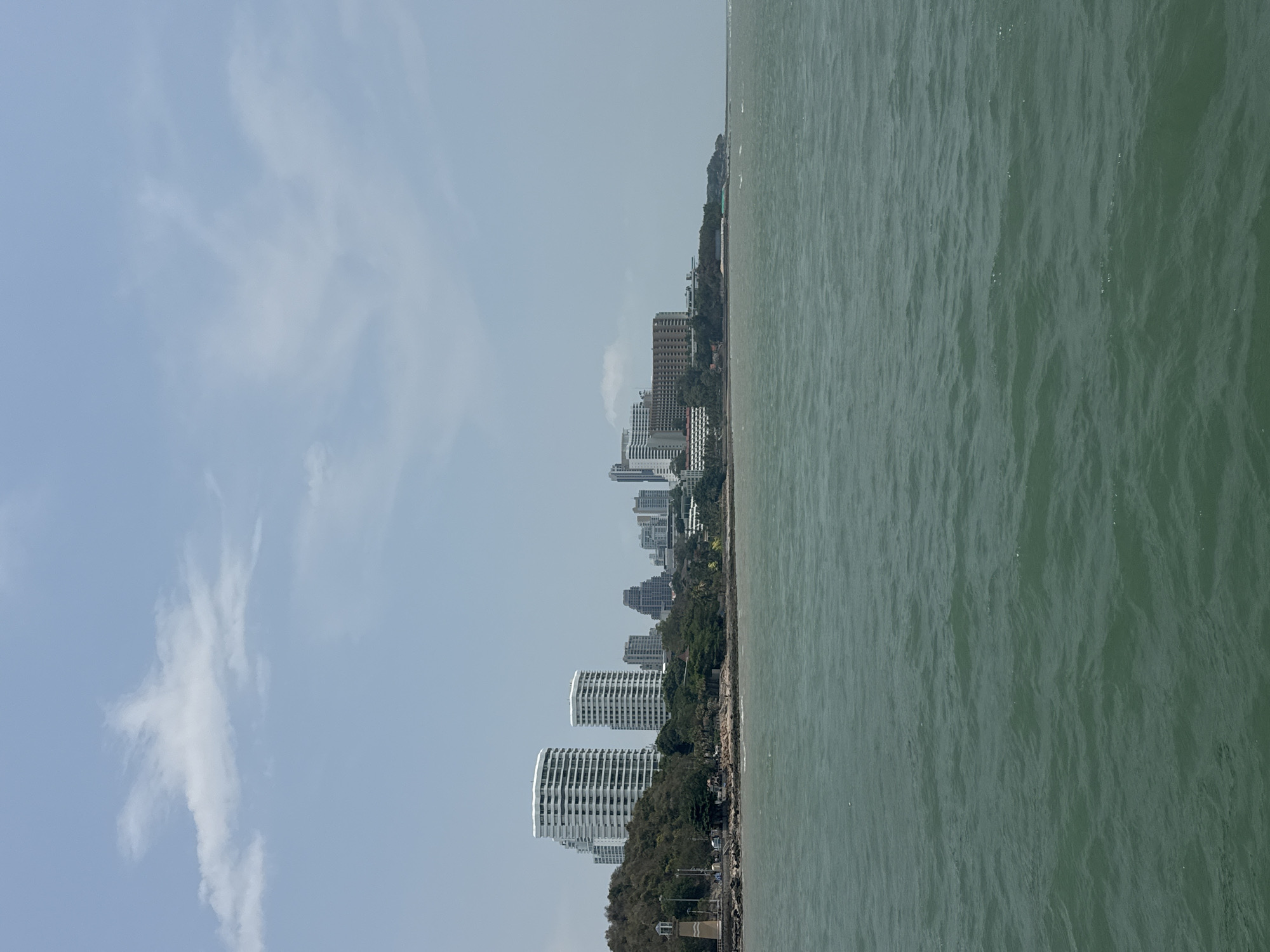 The image depicts the coastline of Pattaya City, Thailand, under a partly cloudy sky. High-rise buildings of varying heights are visible along the shoreline, interspersed with trees. The water is a green hue and stretches to the edge of the frame. The image is taken from a high vantage point. There is no visible human activity in the frame.