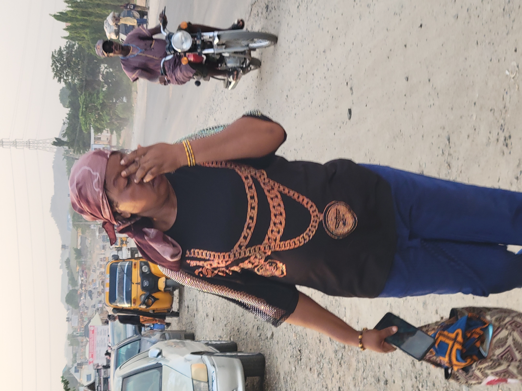 An adult woman wearing a burgundy headscarf, a black t-shirt with a gold-toned chain graphic, blue pants, and gold bracelets stands on a road in Madalla, Nigeria. She holds a black smartphone in her left hand and raises her right hand to her face, appearing to shield her eyes. A patterned bag rests on the ground near her. In the background, a yellow tricycle taxi (keke) and several cars are visible on a congested road with distant buildings and trees. Two men are riding a motorcycle on a road above the woman. The sky appears hazy.