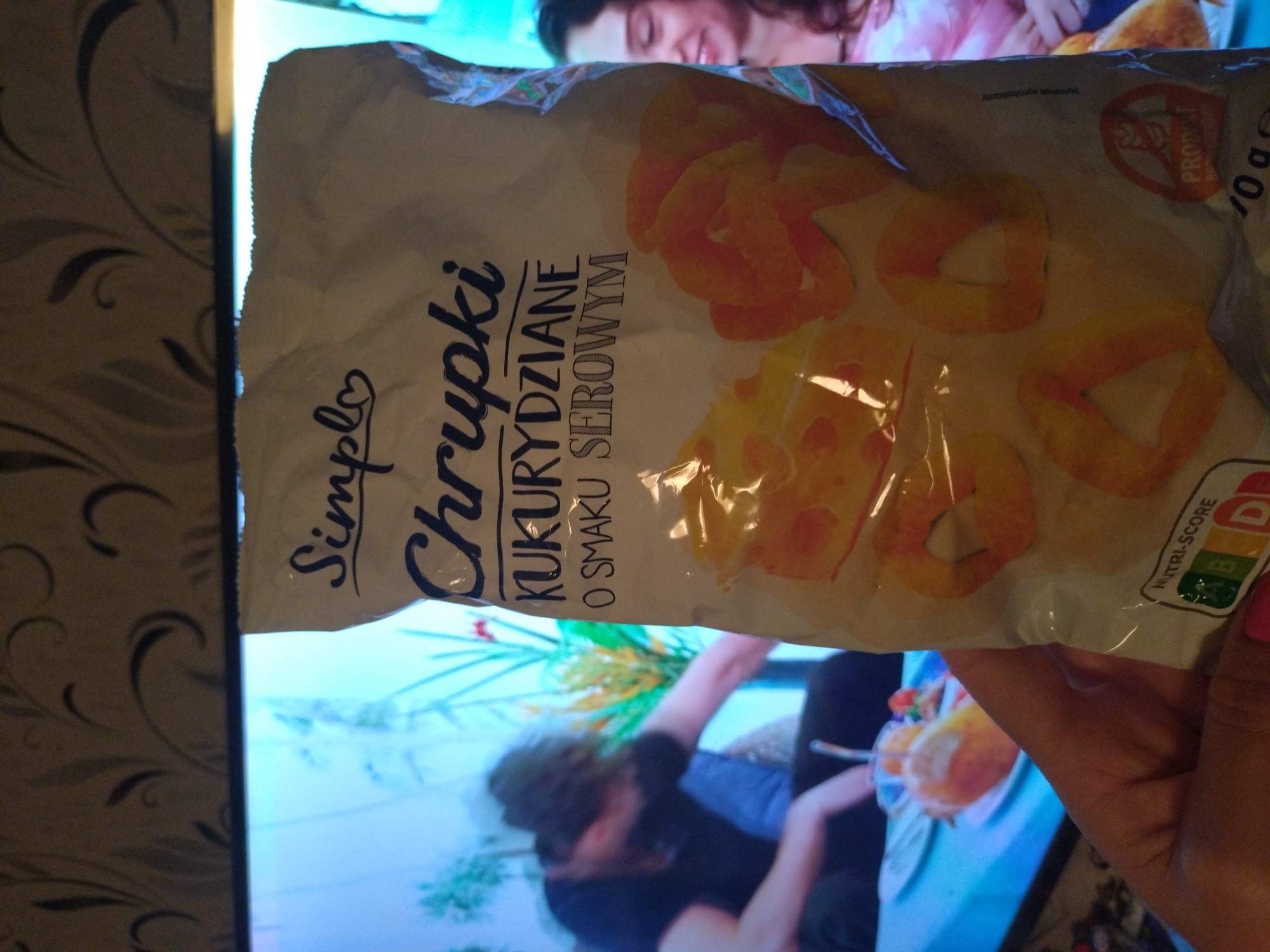 A hand holds a bag of "Simplo Chrupki Kukurydziane O Smaku Serowym" (Simplo Corn Crisps with Cheese Flavor). The bag features images of orange, ring-shaped snacks. The background shows a television screen displaying a scene with people and objects. A wall with a patterned design is visible to the side. The photo does not contain information to suggest it was taken in the city of Stara Zagora, Bulgaria.