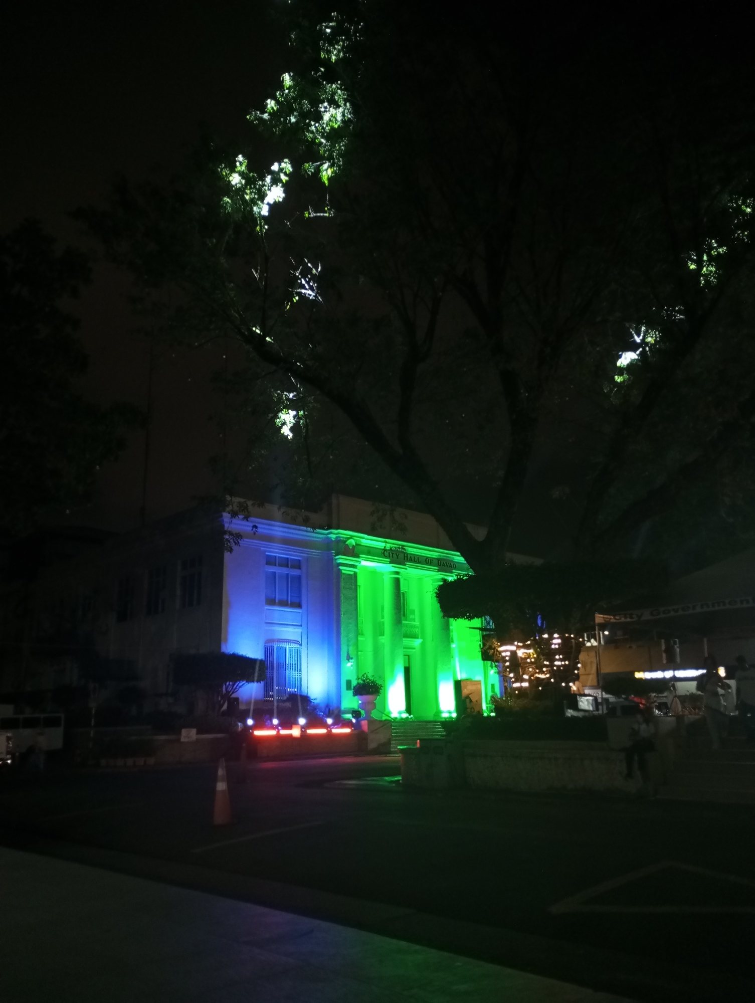 An exterior nighttime shot captures the Davao City Hall illuminated with green and blue lights. A large tree with bright foliage from uplighting dominates the foreground, partially obscuring the building. The architecture of the city hall features classical columns and is lit in vibrant green on the right and blue on the left. Red and pink lights line the street in front of the building. An orange traffic cone is visible on the road. In the background, blurred lights suggest a market or event. Several figures are present but indistinct due to the low light conditions.