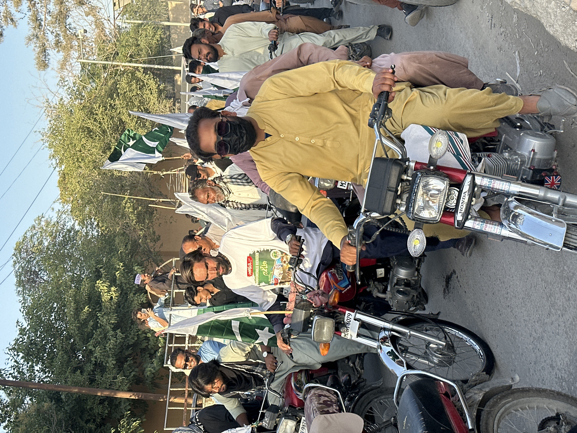 The image shows a gathering of men on motorcycles, possibly in Quetta City Tehsil, Pakistan. They are lined up on a paved surface, likely a street, with trees and buildings visible in the background. The men are wearing a variety of clothing, including traditional Pakistani attire and casual Western clothing. Many of them are holding Pakistani flags. Some are wearing masks. The motorcycles are a mix of models. The presence of Pakistani flags and the organized arrangement of the motorcycles suggests a demonstration or parade. The presence of license plates with registration prefixes for Balochistan Province suggests a location in Quetta City Tehsil, Balochistan, Pakistan.