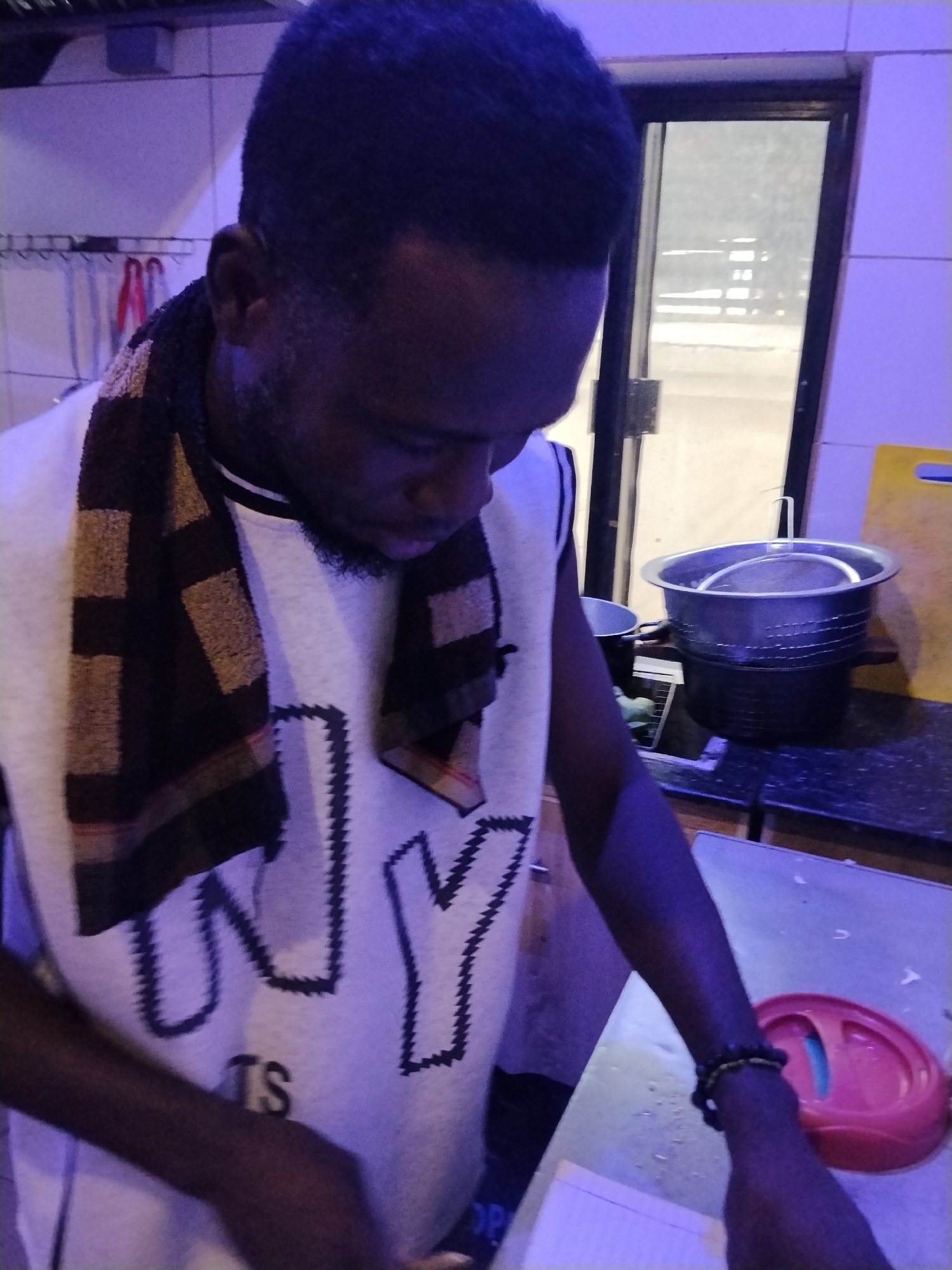 A Black man with dark skin, short hair, and a short beard stands in a room that appears to be a kitchen, possibly in Howayi, Nigeria. He is wearing a white t-shirt with the word "NY" in large black letters and the letters "TS" in smaller black letters below it. A brown and tan patterned scarf is draped around his neck. In the background, there is a stainless steel pot with a strainer inside, a yellow cutting board, and a door. The lighting in the room is dim and cast in a purplish hue, suggesting artificial lighting. The man is looking down at something on the counter in front of him, his hands are not clearly visible.