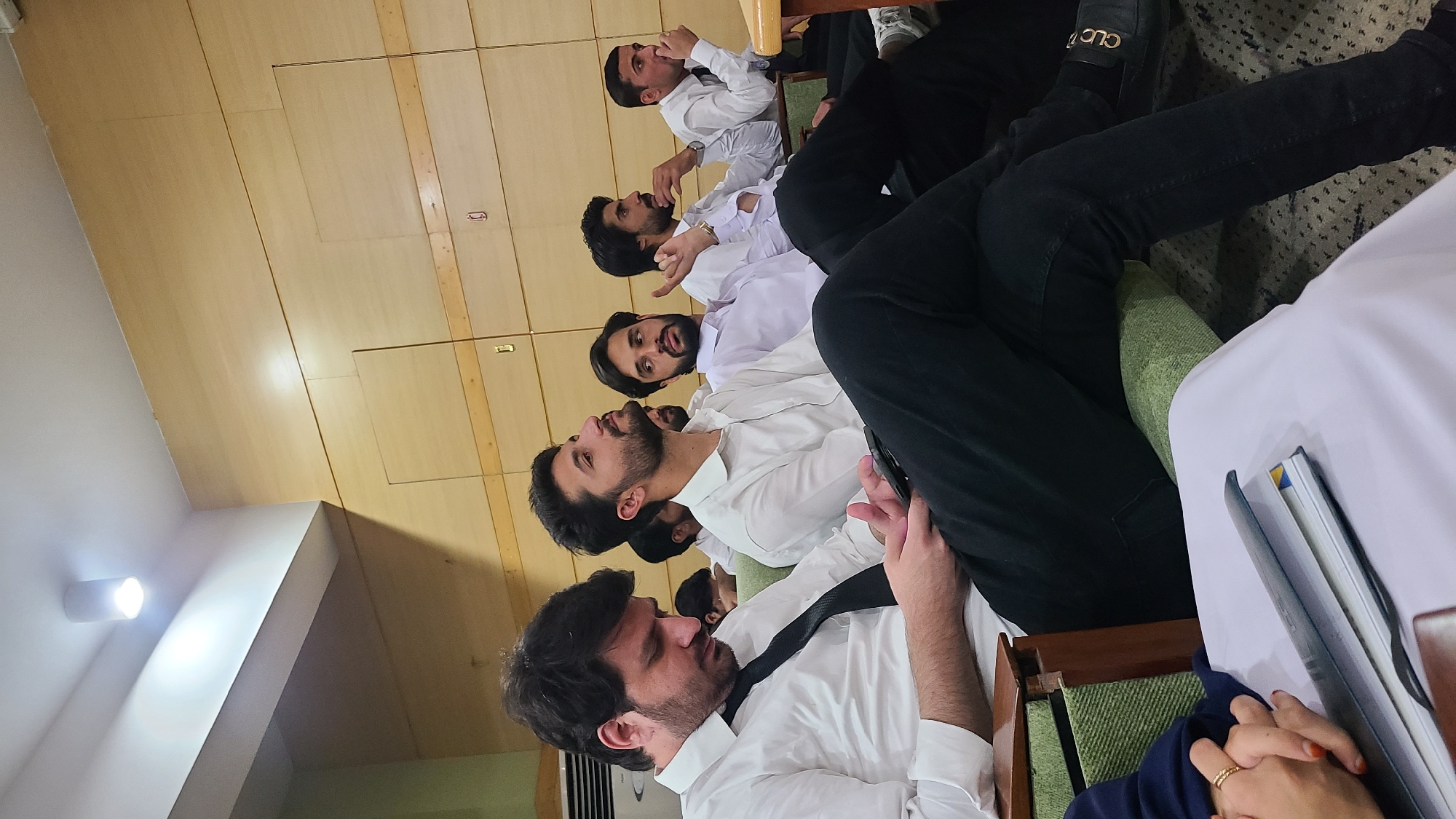 The image captures an indoor gathering of several men, primarily young adults, seated in what appears to be a meeting room or lecture hall in Quetta City Tehsil, Pakistan. The men are mostly wearing white shirts; one is seen with a black tie, while others appear to be in traditional white attire, possibly shalwar kameez. They are generally looking forward or slightly upwards, suggesting they are attentively observing a speaker, presentation, or event.
The setting features light-colored wooden paneled walls or ceiling, with some plain white wall sections. A bright, wall-mounted light fixture illuminates part of the room. The seating consists of chairs with green upholstered backs and seats.
In the foreground, a person's legs are visible, one wearing black pants and black loafers with "GUCCI" branding, and another with a light green sock. Also visible are hands, possibly belonging to another attendee, holding a book or folder, adorned with rings and showing manicured nails. The attire and attentive postures indicate a formal or semi-formal educational or professional event. The specific time of day is not discernible due to the indoor setting and artificial lighting.