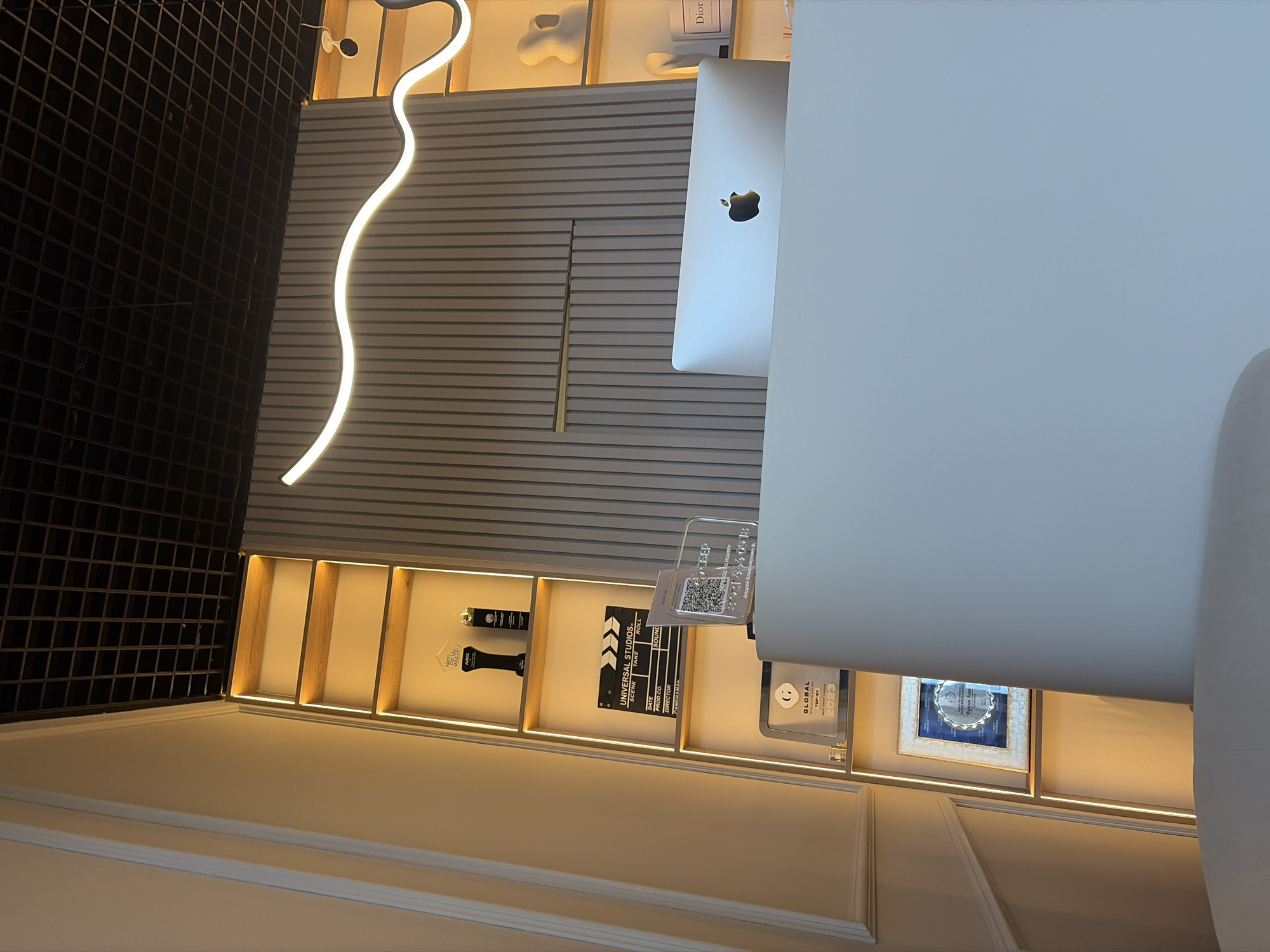 Captured from an overhead perspective, the image showcases a modern office setting, likely situated in Saint Petersburg, Russia. The scene is dominated by an Apple iMac positioned on a white desk, accompanied by a white office chair. The wall behind the desk features horizontal paneling and open shelving. These shelves hold various decorative items, including a Dior book, awards, a movie clapperboard labeled "UNIVERSAL STUDIOS," and a framed picture. The room is illuminated by a decorative wavy light fixture and the integrated lighting within the shelves. The overall atmosphere is contemporary and professional.
