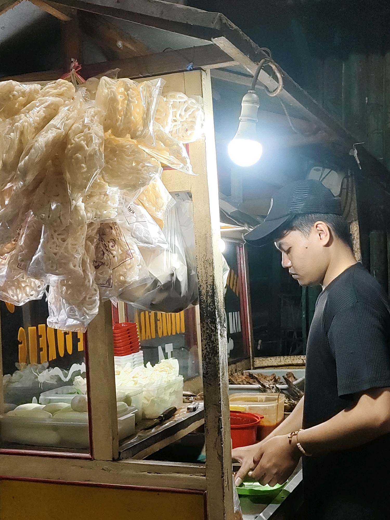 A young man wearing a black t-shirt and a black baseball cap is standing at a food stall at night in Probolinggo, Indonesia. He is looking down and appears to be preparing food. The stall is brightly lit by a single overhead bulb. Various food items are visible, including bags of crackers hanging from the ceiling and containers of prepared ingredients on the counter. The stall itself is made of wood and metal, with a glass display case in the front. The background is dark, suggesting it is nighttime.