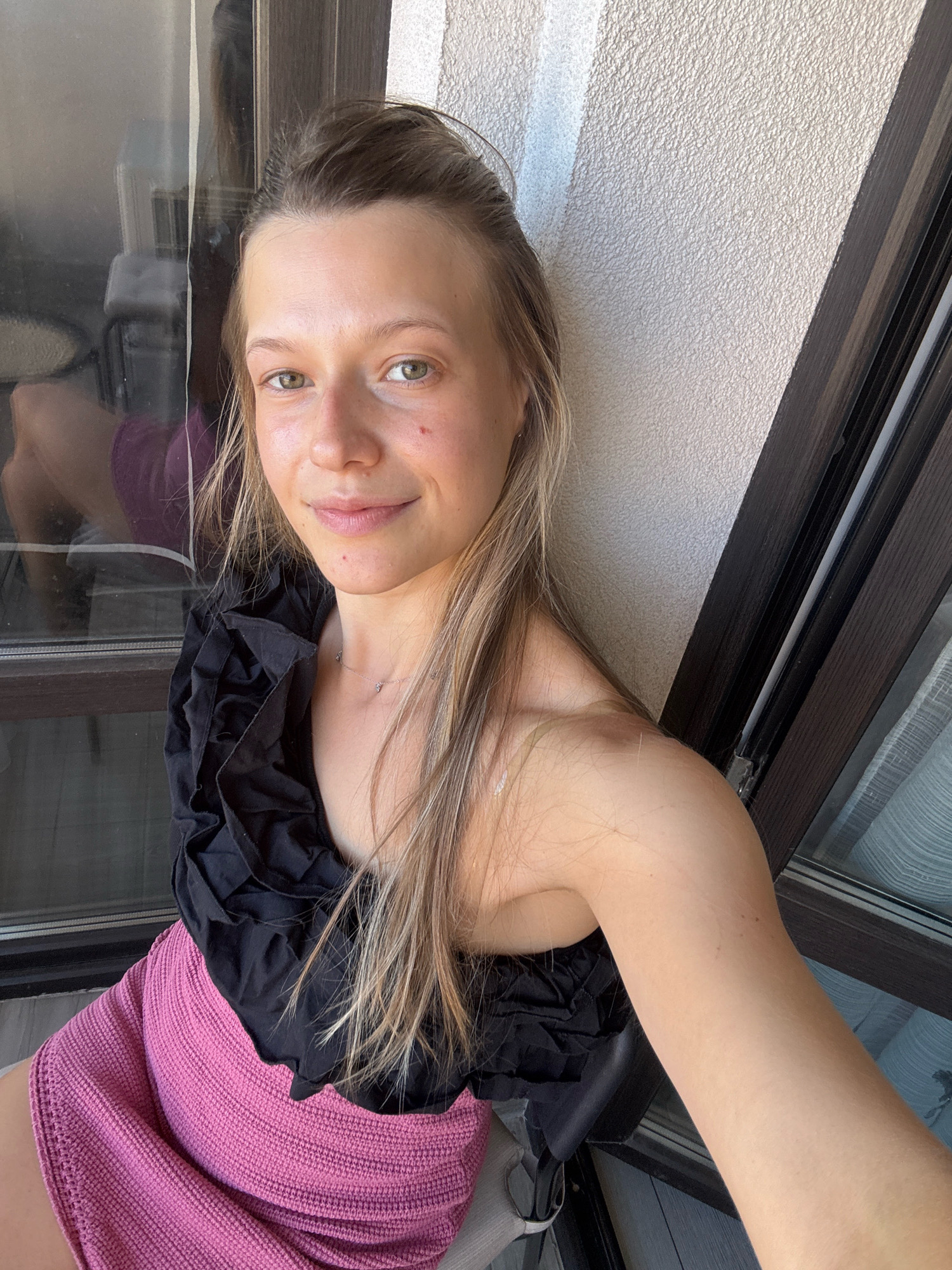 A young woman with blonde hair pulled back and a black ruffled top and pink skirt takes a selfie. She is sitting on a balcony with a tiled floor. Behind her, a sliding glass door reflects an interior space with furniture. The wall behind her is textured and painted white. The time appears to be daytime. The location is not specified in the image or metadata.
