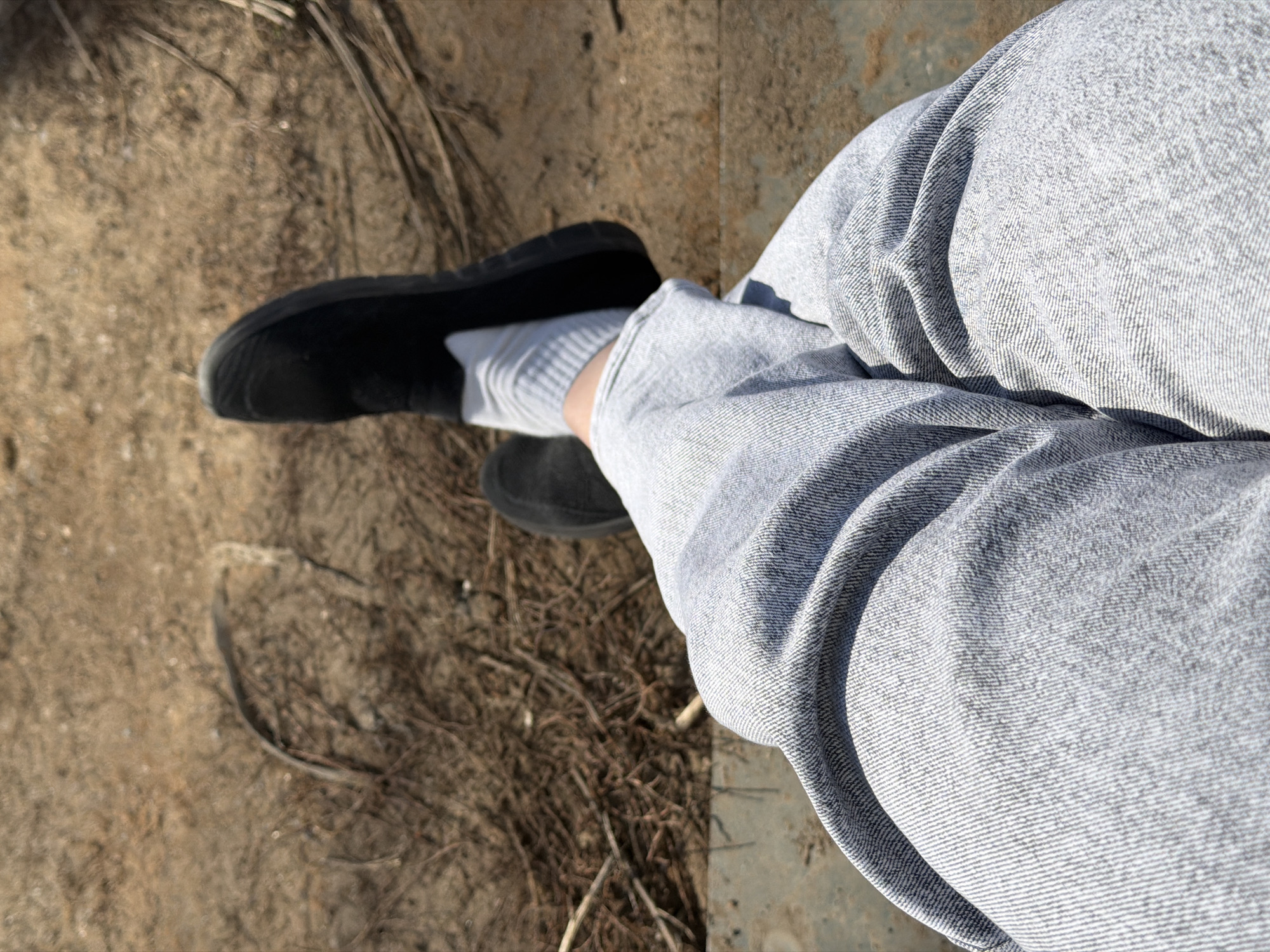 The image shows a person sitting on a metal surface, with their legs bent at the knees and positioned to the right side of the frame. They are wearing light gray denim jeans, white socks, and black shoes. The background consists of dry dirt and twigs. The location is not mentioned in the image.