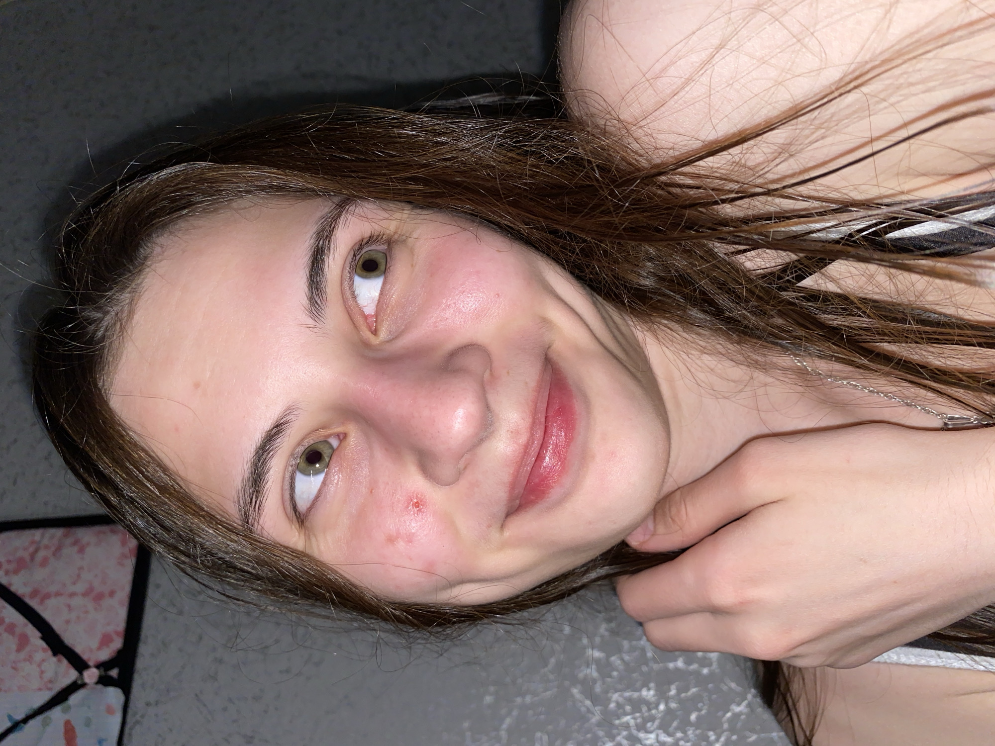 A photograph shows a young woman with light skin, brown hair, and hazel eyes. She is smiling slightly. There is a blemish on her left cheek. She is wearing a striped top and a silver necklace. Her hand is near her neck, touching her hair. The background is a gray surface. There is no discernible location visible in the image, and therefore no information to determine a location in Votkinsk, Russia.