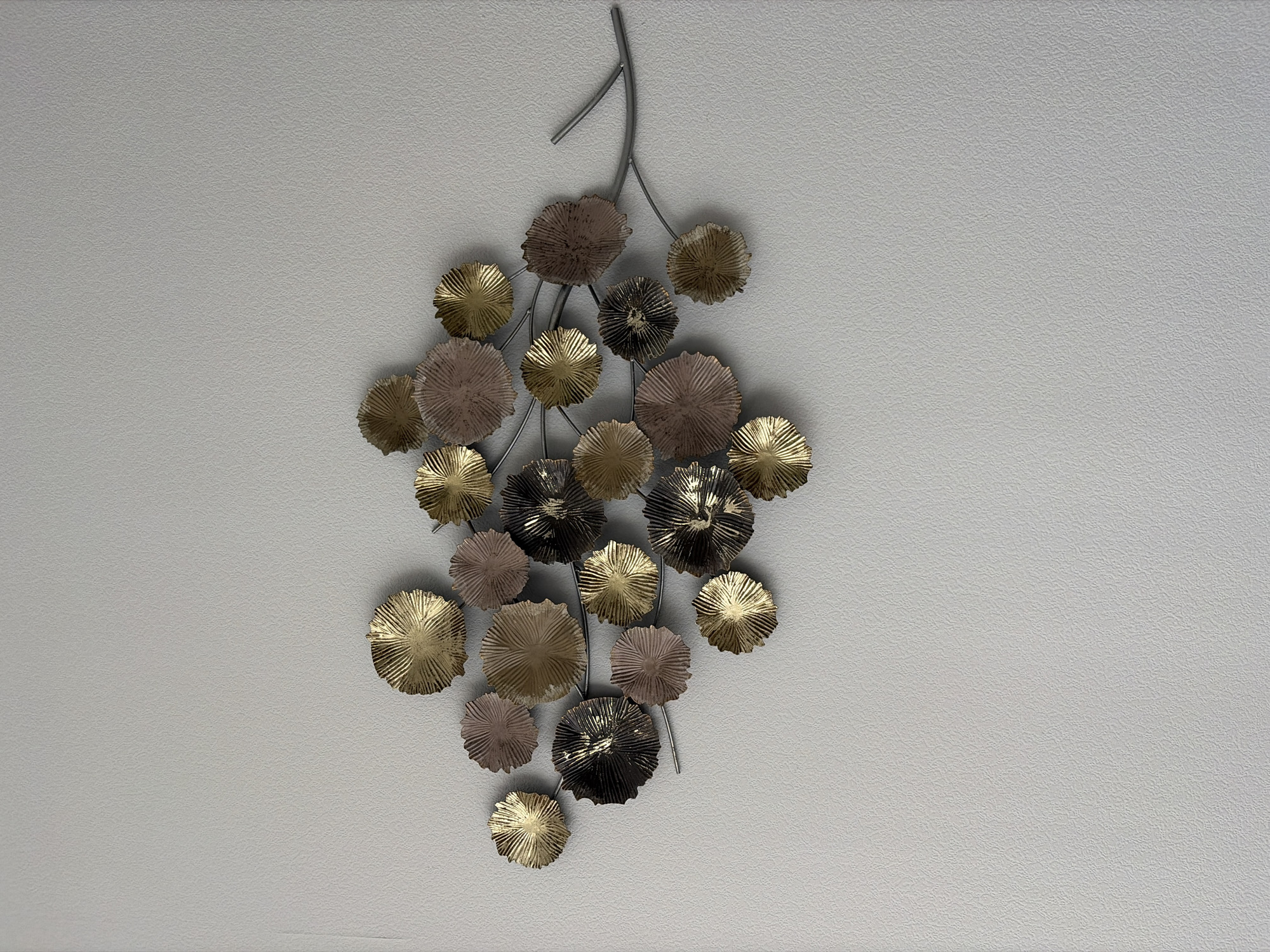 The image shows a metallic wall art piece mounted on a textured light gray wall. The artwork is shaped like a branch with numerous round, textured leaf-like or petal-like elements attached to it. These elements vary in color, appearing in shades of gold, a muted rose gold or blush, and a dark metallic color, possibly black or dark bronze with a metallic sheen. The elements are clustered together, creating a visually dynamic arrangement that resembles a flowering branch or a cluster of stylized leaves. The branch itself is thin and metallic, extending horizontally across the frame. The lighting suggests the image was taken indoors, likely in a domestic setting, but no specific location in Kotelniki, Russia, can be determined from the visual information provided.