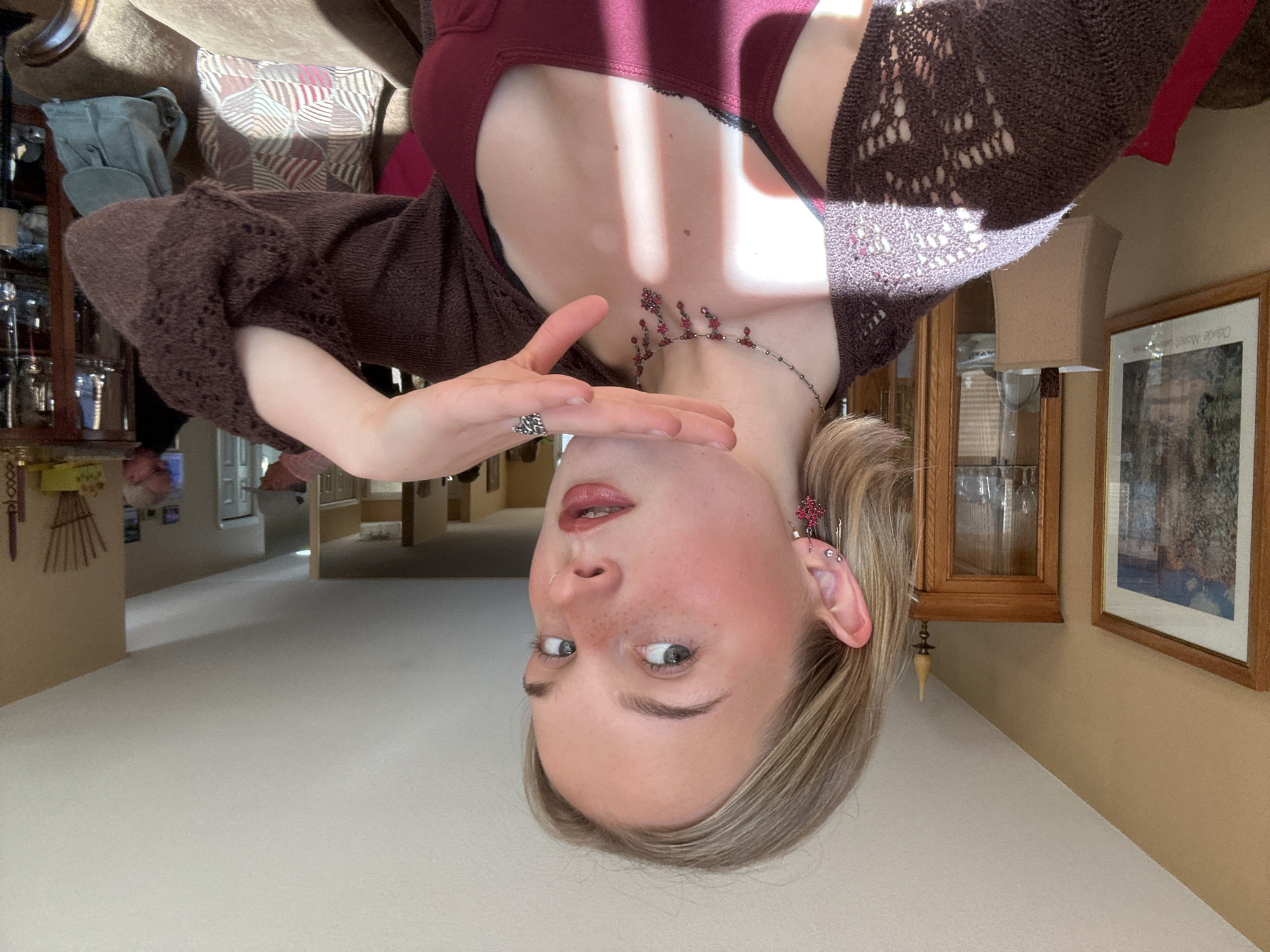 A fair-skinned person with blonde hair and blue eyes is positioned upside down in the foreground. She is wearing a burgundy tank top and a brown crocheted sweater. She has a red and silver necklace and a silver ring on her left hand, which is raised towards her face. Her ear is adorned with multiple earrings. The background includes a living room setting with a couch, a glass cabinet, a wooden wall clock, and framed artwork. The scene appears to be indoors during daylight hours. No specific indicators confirm this is in Kelowna, Canada.