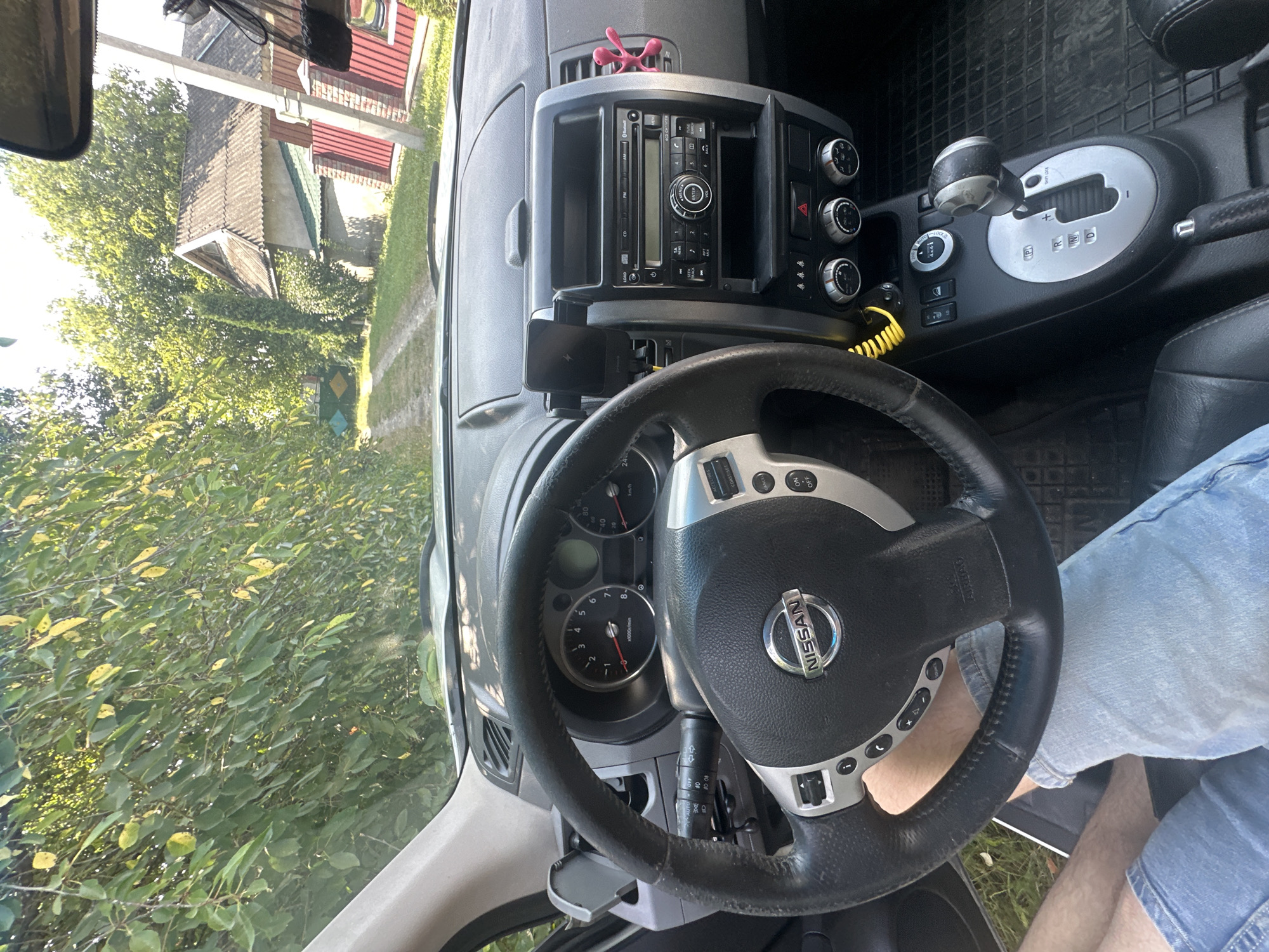 The image captures the interior of a Nissan X-Trail, viewed from the driver's perspective. The dashboard features a steering wheel with the Nissan logo, instrument cluster displaying speed and RPM, and a central console with a car stereo and climate controls. A mobile phone is mounted on the dashboard. Outside the car, visible through the windshield and side windows, is a rural setting with trees, a dirt road, and a house with a red fence, suggestive of a location in Shatava, Ukraine. The driver's legs, clad in denim shorts, are visible in the lower part of the frame, indicating the car is stationary.