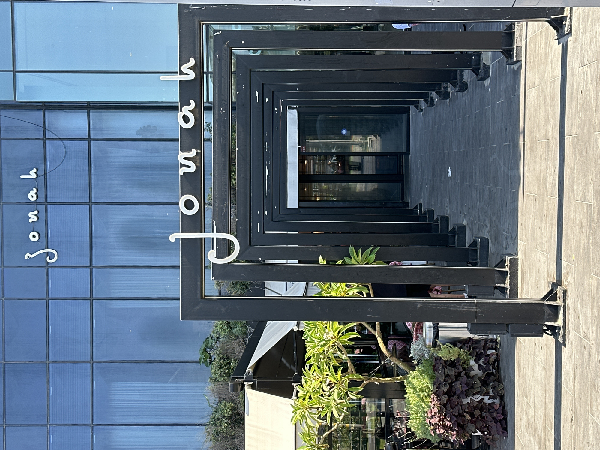 The image shows the entrance to a building with "Jonah" written in white cursive letters on the facade, and again in similar lettering on an overhead structure framing the walkway. The building features a large glass facade on the upper floors, reflecting the sky and surrounding structures. A pathway, paved with stone tiles, leads into the building, flanked by a series of black metal rectangular frames. To the left of the walkway, there are planters with green and purple foliage, and a tree with bright green leaves. The scene appears to be outdoors, likely in a public or commercial area. The location is identified as Netanya, Israel. No people are clearly visible or engaged in any distinct actions, though there are reflections and faint figures in the background.