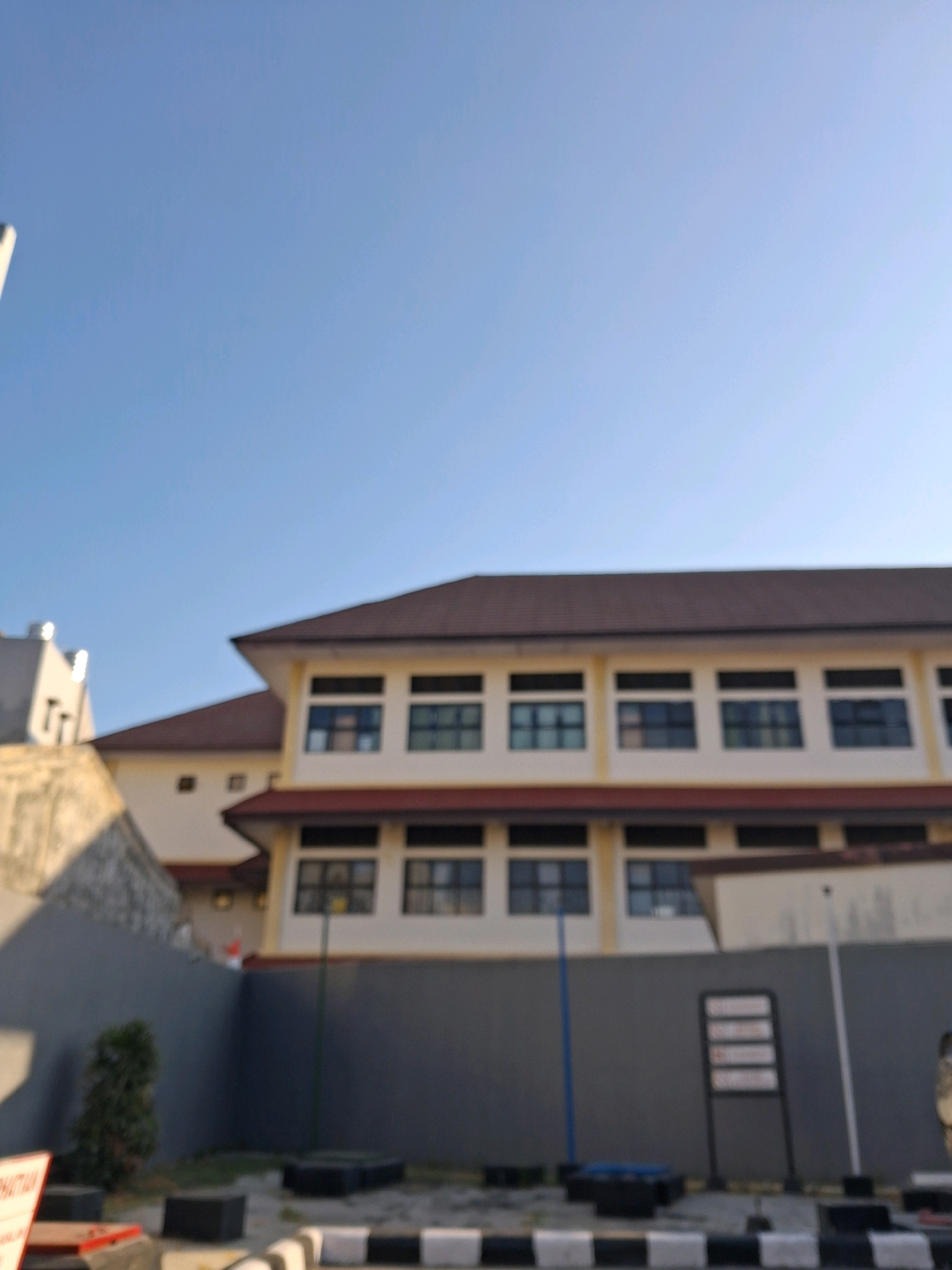 A low-angle, out-of-focus exterior shot shows a modern, light-colored building with a brown roof and multiple rows of dark windows under a clear blue sky. The building appears to be two stories tall, with a gray wall extending horizontally in front of the lower level, partially obscuring the lower windows. A small, dark green bush is visible behind the gray wall. To the left of the building, a portion of another structure, also light-colored, is visible, with a textured, light-brown wall in the foreground. The ground in front of the gray wall has black and white striped markings and several black, rectangular objects. A blue pole stands near the building and to the right, a black stand with multiple white rectangular signs is visible. The scene appears to be an institutional or educational building in Surakarta, Indonesia.