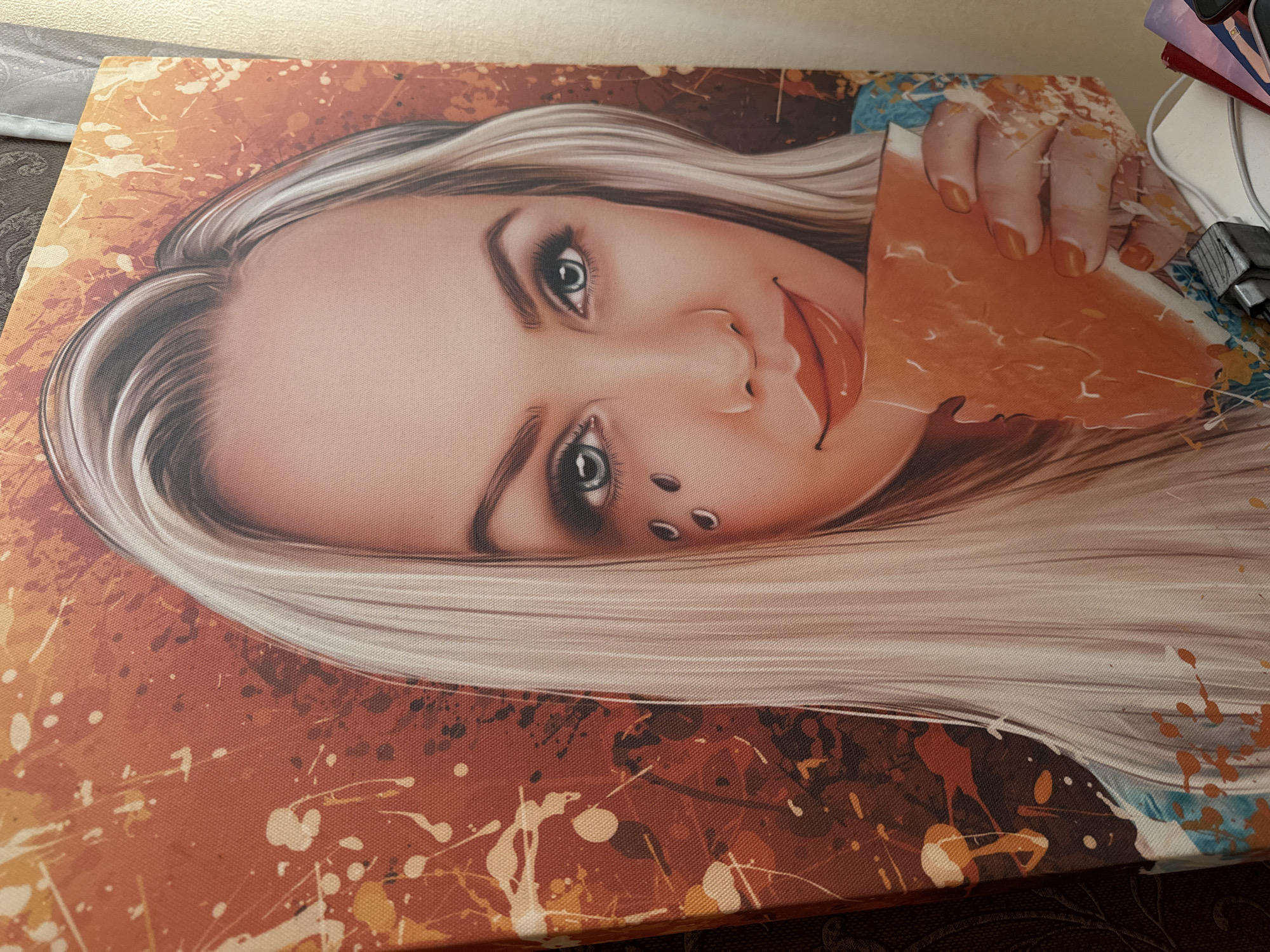 The image displays a close-up photograph of a vibrant, stylized portrait printed on a stretched canvas. The canvas is positioned on a dark, textured surface, possibly a table or desk, with part of a lighter wall or surface visible in the upper background.
The central subject of the artwork is a young woman's face, tilted clockwise. She has long, straight, light blonde or silver hair. Her eyes are open, with prominent eyelashes and defined eyeliner, and appear to be blue or teal. She has full, reddish lips and two metallic piercings on her left cheek, below her eye. Her left hand, with orange-painted fingernails, is visible near her mouth, holding a torn, irregularly shaped piece of orange material that resembles paper or fabric.
The background of the portrait is an abstract, energetic splatter pattern, featuring various shades of orange, brown, and white paint drips and splashes. Hints of blue and teal splatters are visible on the far right side of the canvas. The texture of the canvas weave is clearly visible throughout the image, giving it a tangible quality. In the upper right corner of the real-world scene, several objects are partially visible, including a white power cord, a black electronic device, and some stacked items with colorful spines. There is no visible text in the image.