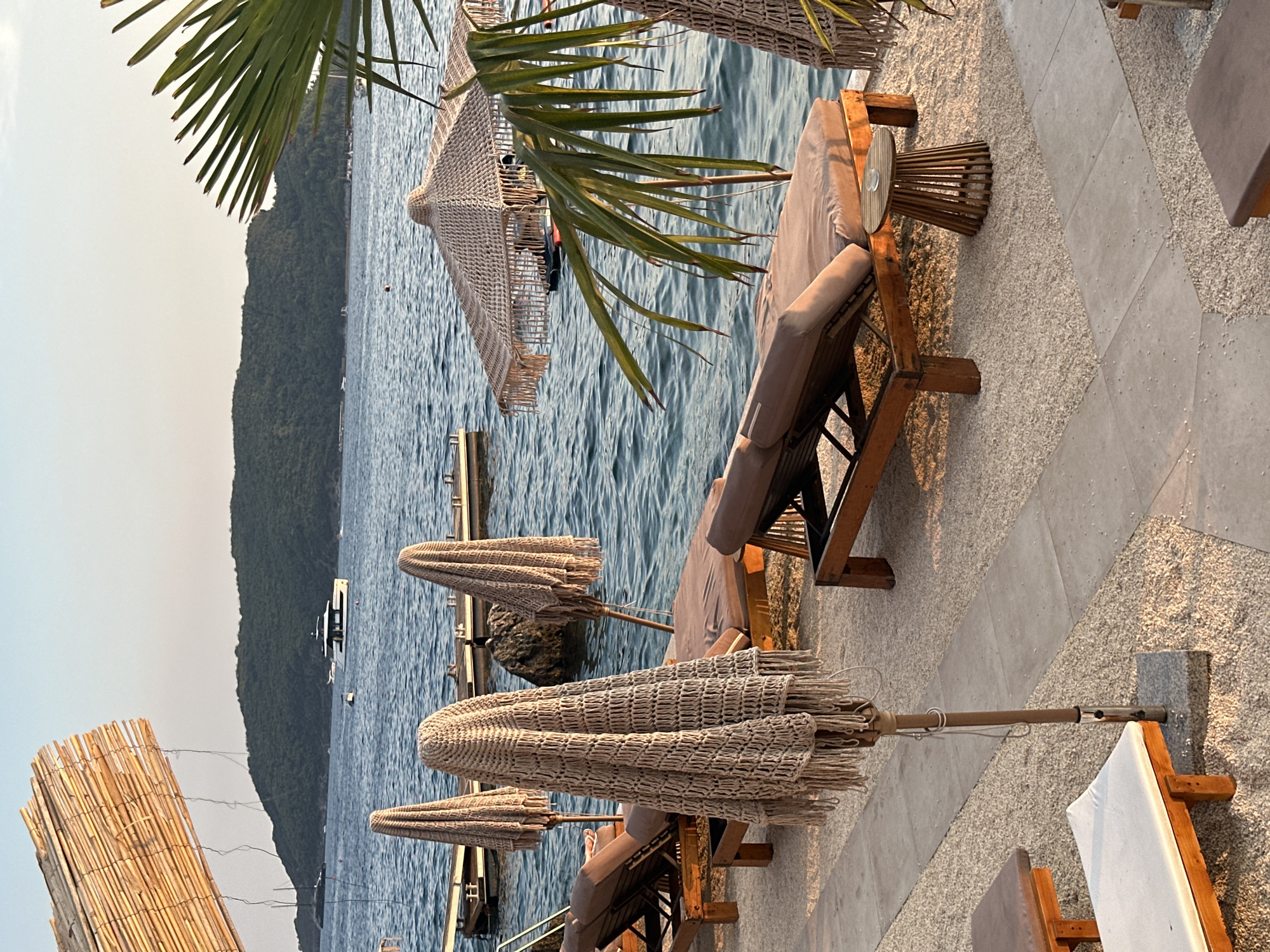 This image depicts a tranquil beachside scene at sunset in Budva, Montenegro, likely on the popular Mogren Beach or a similar coastal resort. In the foreground, several sun loungers are arranged on a gravelly ground, some with beige cushions. Two of the loungers are partially visible in the lower portion of the image, one with a white cushion. Behind these, a row of wooden-framed sun loungers with beige cushions stretches across the mid-ground, each accompanied by a decorative, tasseled beach umbrella that is closed. One of these umbrellas is intricately crocheted. A small, cylindrical wooden side table with a glass on top is situated between two of the loungers.
The water of the Adriatic Sea is calm and reflects the soft light of the setting sun, with gentle ripples across its surface. In the background, a lush green, mountainous island dominates the horizon. Several boats, including a sleek white yacht, are anchored or cruising on the water, adding to the sense of a popular seaside destination. To the far left, a bamboo-like sunshade structure extends into the frame. A large palm frond from a nearby tree drapes over the right side of the image, adding a tropical element to the scenery. The overall atmosphere is serene and inviting, suggesting a relaxed holiday setting.