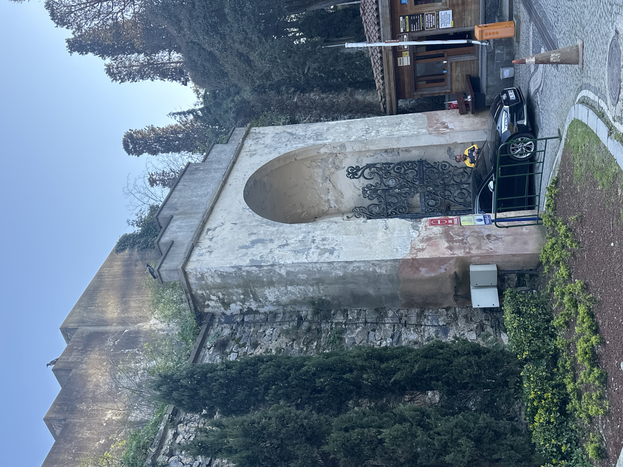 The image shows a historical stone structure in Beşiktaş, Turkey. The structure features an arched opening with a decorative iron gate. A black car is parked in front of the gate, with a person visible inside wearing a yellow shirt. Next to the car is a small booth or building with signage. A traffic cone is placed near the vehicle. The area around the structure is landscaped with trees and greenery. Stone walls and other architectural elements are visible in the background, with a clear sky above.