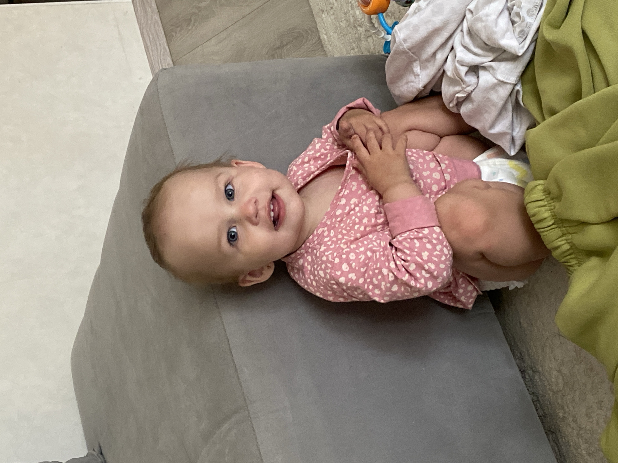 A baby with blue eyes and blonde hair is sitting on the floor next to a gray couch. The baby is wearing a pink shirt with a white pattern and a diaper. There are some clothes on the floor in front of the baby, including a green garment and a white cloth. The background shows a wall and wooden flooring. The scene appears to be in a residential setting in Chaikovsky, Russia.