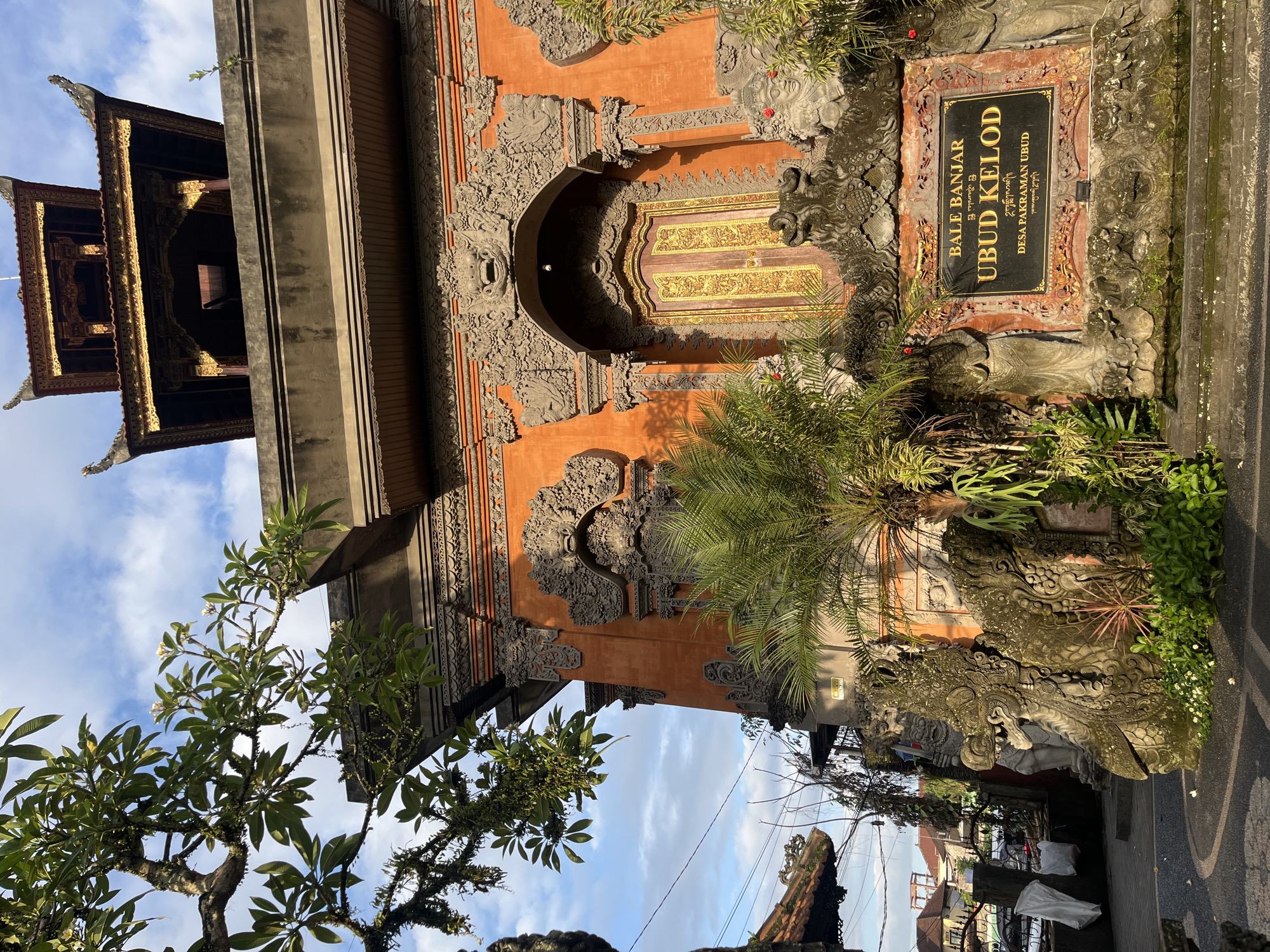 The image depicts the entrance gate to Bale Banjar Ubud Kelod in Ubud District, Indonesia. The gate is intricately decorated with carved stone details and painted in shades of orange. A wooden tower-like structure is visible on the left side. Lush greenery surrounds the gate, including a tree on the left side and a small palm tree in front. A sign on the right side identifies the location as Bale Banjar Ubud Kelod, Desa Pakraman Ubud. The sky is visible in the background, showing a mix of blue sky and white clouds.