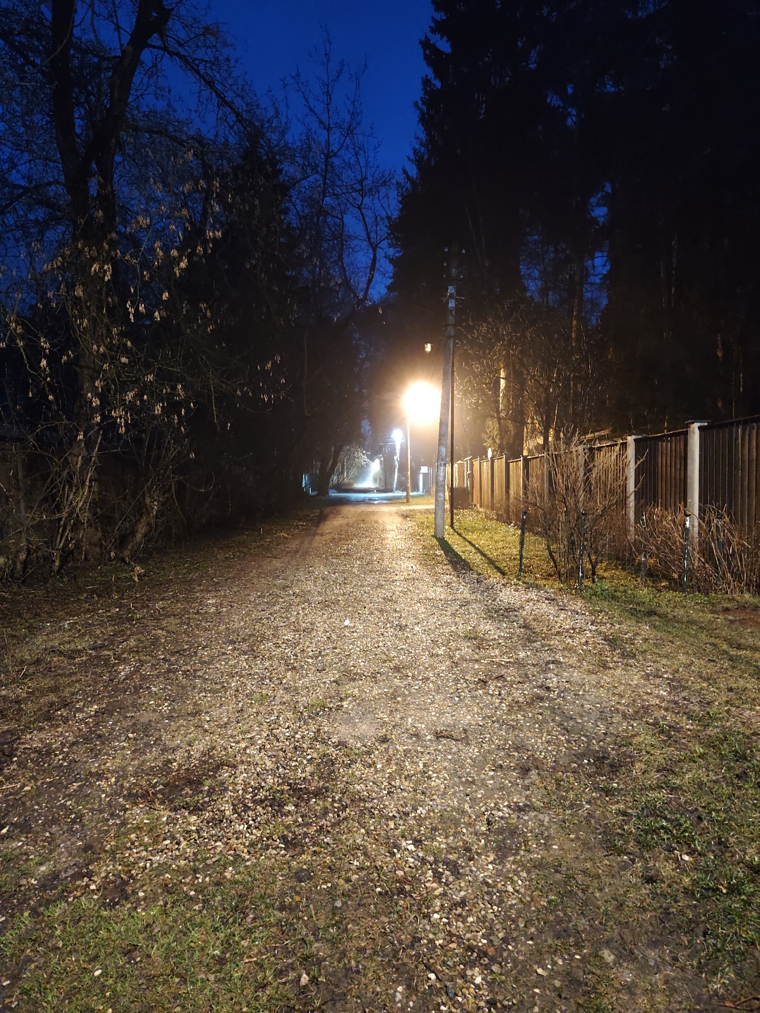 The image depicts a dimly lit path at night, likely in the city of Kratovo, Russia. The path is covered in gravel and lined with grass and vegetation on either side. To the right of the path is a wooden fence. A street lamp illuminates the path, casting a glow on the immediate surroundings. The sky is dark blue, and trees are visible in the background, their branches bare. The overall scene suggests a quiet and secluded outdoor location. There is no visible evidence of people in the image.