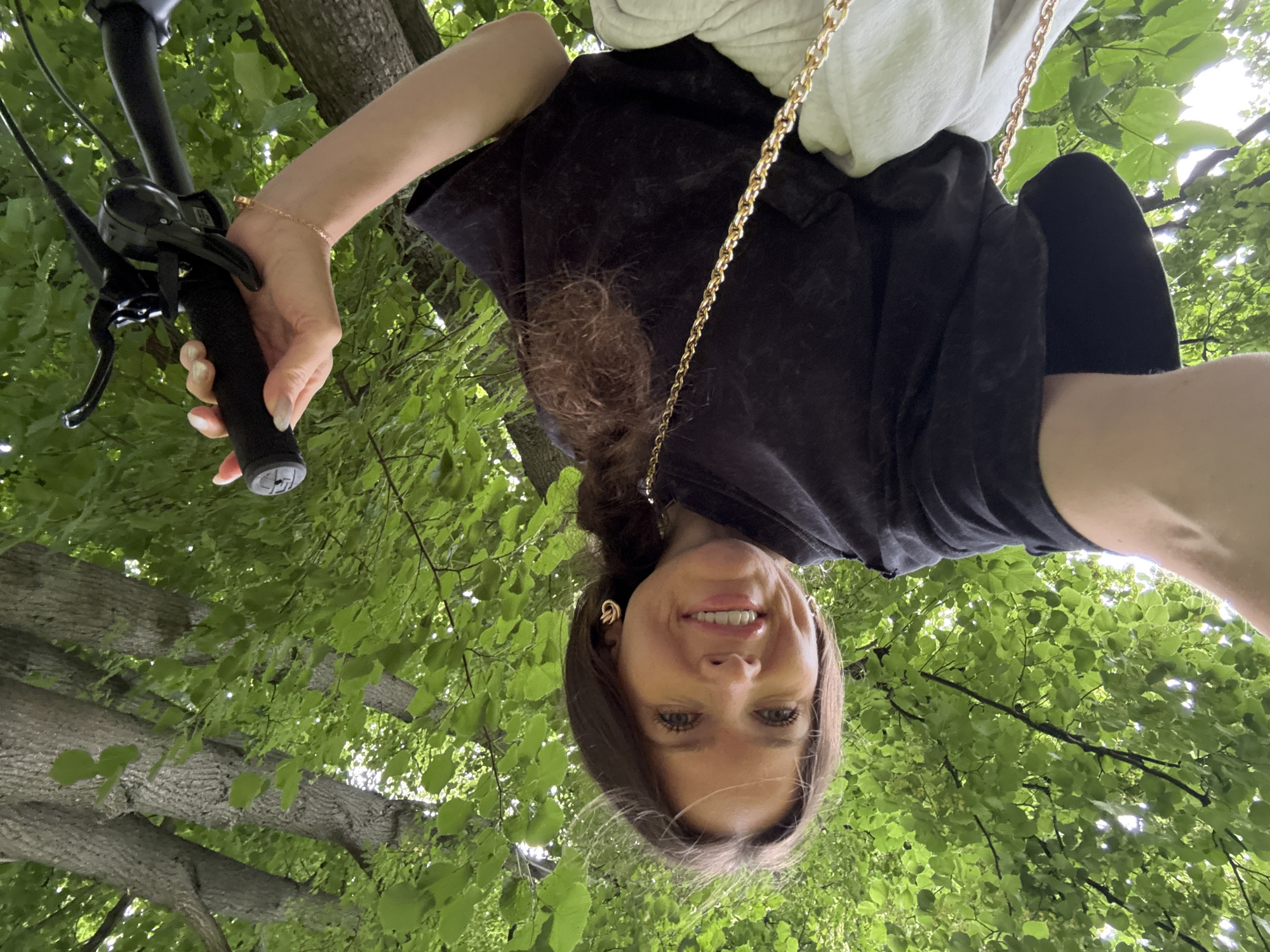 A woman is holding the handlebars of a bicycle, looking up at the camera with a smile. She is wearing a black t-shirt and a light-colored hooded sweatshirt over it. A gold chain is visible draped over her shoulder. She has on gold hoop earrings and a delicate bracelet on her wrist. Her hair is brown and pulled back. The shot is taken from a low angle, looking up through green leaves and tree branches. The sunlight filters through the foliage, suggesting a daytime scene. The overall impression is one of enjoyment and engagement with the outdoors. The image appears to have been taken in Moscow, Russia, given the location context. There is no visible text in the image.