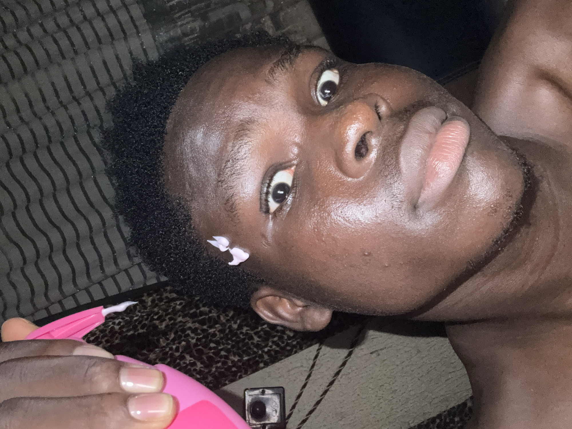 A close-up, low-angle shot depicts a dark-skinned person lying on a patterned surface, possibly a bed or floor. The individual's face is prominent, with eyes wide and lips slightly parted. A small, light-colored object, possibly a flower petal, rests near their left temple. The person is holding a pink object, possibly a beauty tool, in their left hand, with fingernails visible. A small, black object, possibly a camera, is visible near the lower left. There is no information available to determine if this image was taken in the city of Onitsha, Nigeria, or elsewhere.