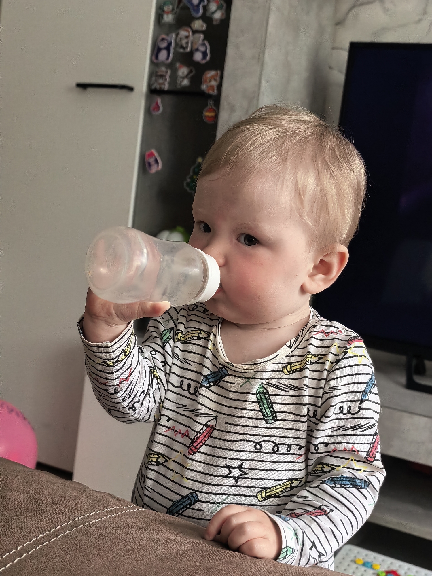 An indoor, eye-level shot captures a blonde-haired baby or toddler drinking from a clear plastic bottle. The child, with rosy cheeks and a focused expression, is dressed in a long-sleeved onesie featuring horizontal black stripes and colorful crayon drawings. They hold the bottle with both hands, positioned to their mouth, appearing to drink a clear liquid.
The setting is a domestic space, likely a living room or common area in Sterlitamak, Russia. In the immediate foreground, a brown, textured sofa or upholstered furniture piece is visible, partially obscuring the lower half of the child. To the left of the child is a plain white wall. The background on the right reveals a dark-colored refrigerator or cabinet door adorned with an assortment of small, colorful magnets, some resembling cartoon characters or festive designs. A portion of a dark-screened television is also visible behind the child's head. Further to the left, a pink balloon is partially visible, while colorful, small toy elements can be seen on the floor to the bottom right.
The lighting is soft and natural, suggesting daytime. The overall scene conveys a quiet, everyday moment of a child hydrating in a comfortable home environment. No discernible text is visible in the image.