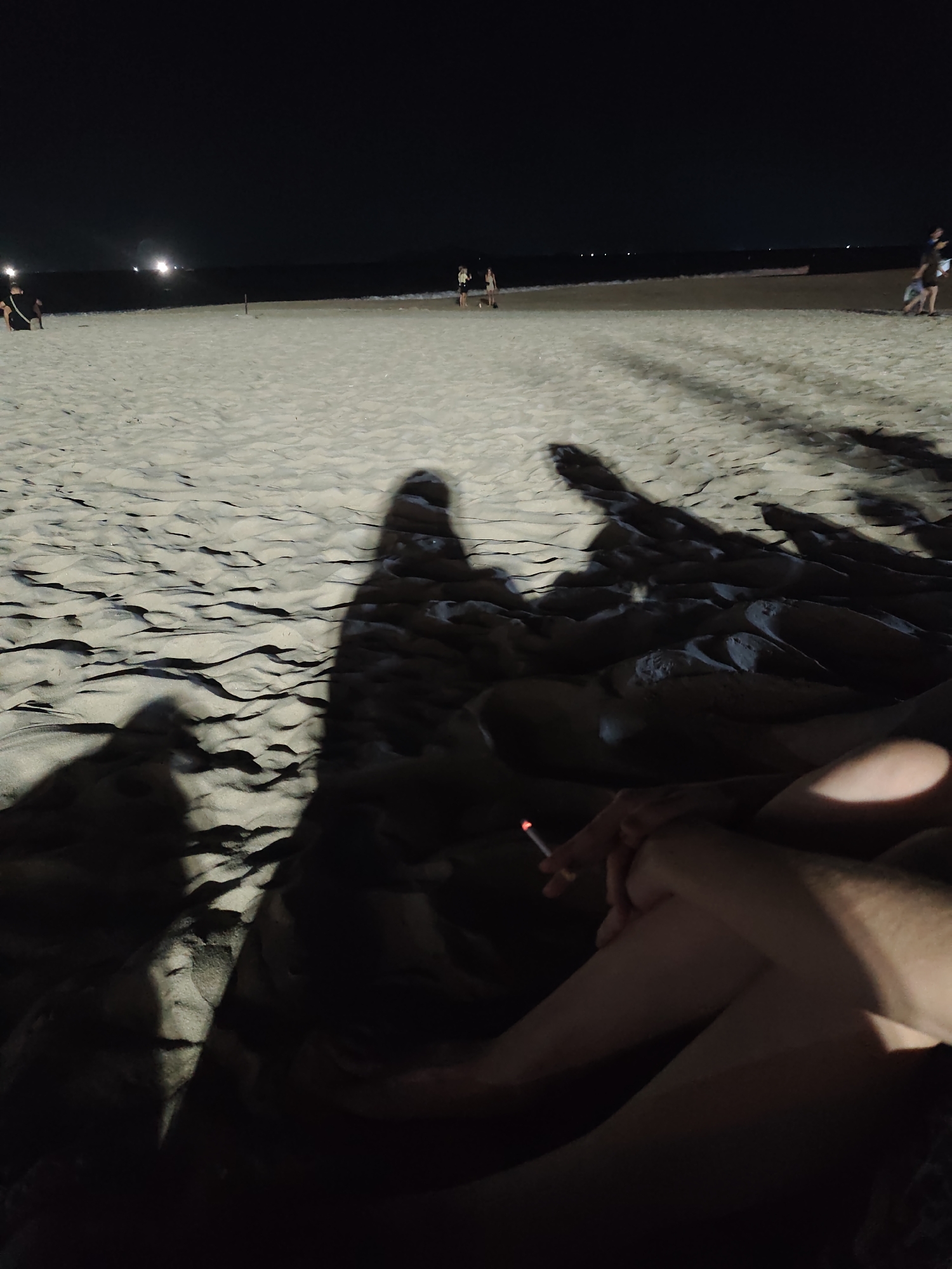 The image depicts a beach scene at night. The foreground is dominated by sand dunes and the shadows cast by people. In the mid-ground, a few individuals are visible, silhouetted against the dark sky and the faint glow of lights in the distance. The sea is a dark expanse in the background, with gentle waves breaking on the shore. The overall atmosphere is serene and tranquil, with the soft glow of distant lights and the vastness of the night sky creating a sense of peace.