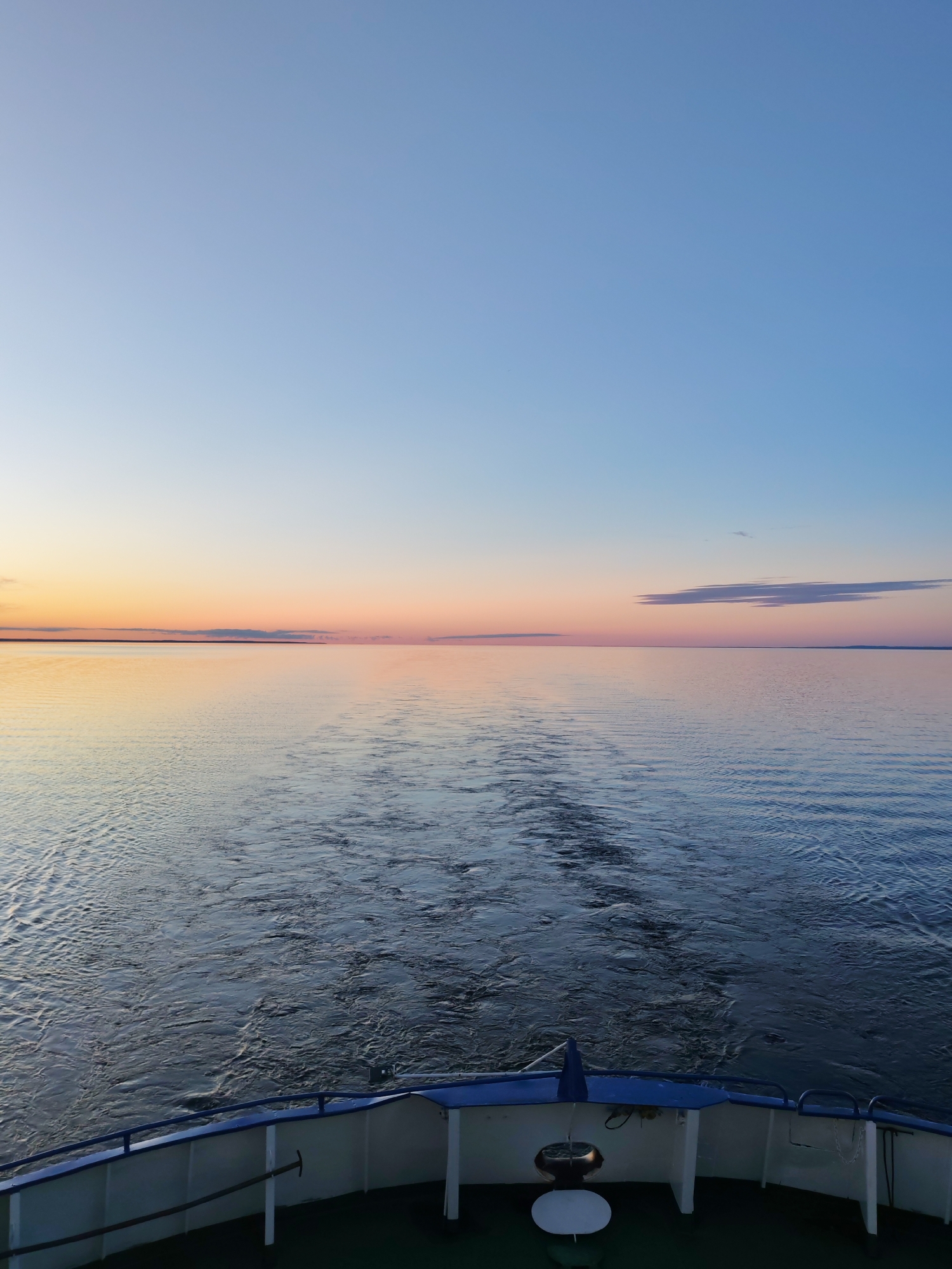 The image captures a serene view from the stern of a boat moving across a vast, calm body of water, likely a large lake or sea, during either sunrise or sunset. The sky displays a beautiful gradient of colors, transitioning from a clear light blue at the top to soft pinks and oranges near the horizon, indicating the golden hour. A few elongated, wispy clouds are visible, with one more prominent cloud stretching horizontally on the right side, reflecting the warm hues of the sky.
The water surface is remarkably still, acting as a mirror that perfectly reflects the vibrant colors of the sky. The boat's movement is evident from the distinct, frothy wake trailing behind it, breaking the reflection and showing the disturbed water.
In the foreground, the stern of a boat is visible. It features a light-colored hull with a blue railing along the top edge, suggesting a passenger vessel. Parts of the boat's deck, railings, and what appears to be some equipment or seating are visible. The perspective is looking directly back from the boat, emphasizing the endless horizon and the boat's path.
The overall atmosphere is one of tranquility and vastness. There is no visible text in the image. The location is noted as Unknown, Russia.