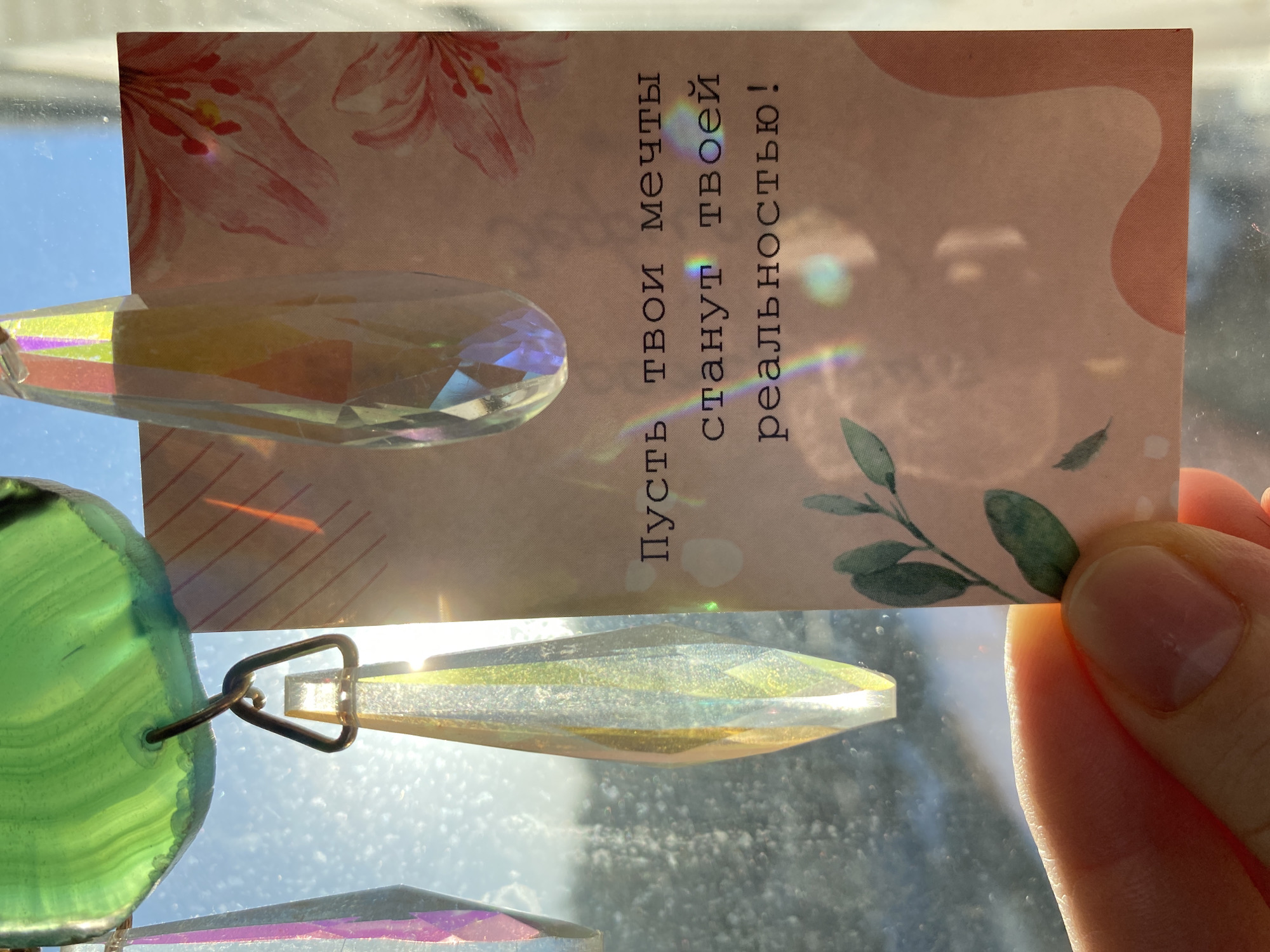 A person holds a small card with pink floral patterns and the text "Пусть твои мечты станут твоей реальностью!" in front of a suncatcher hanging in what appears to be a window. The suncatcher has green and crystal-like elements that reflect light. The background shows a bright, possibly outdoor scene, suggesting a sunny day. It is difficult to confirm the location with certainty, but based on provided information, it is in Chaikovsky, Russia.