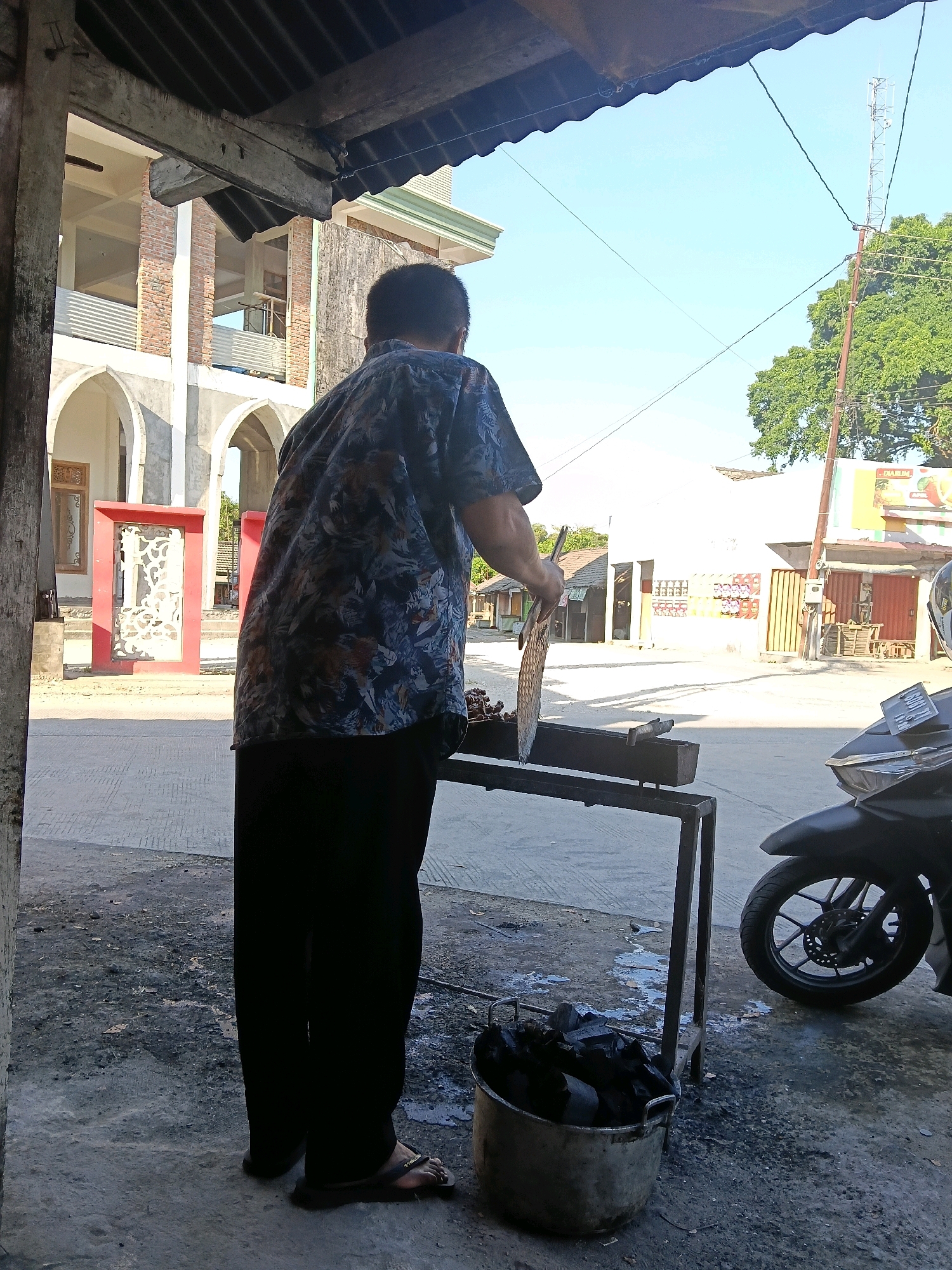 The image depicts an outdoor scene in the city of Kedungtuban, Indonesia. A person, viewed from the back, is standing and appears to be grilling. They are wearing a patterned short-sleeved shirt and dark trousers. In front of them is a metal grill with what looks like meat on it. To the left of the grill, there is a metal bucket filled with charcoal. The ground is paved, and there is a building with arched windows in the background, along with other structures that suggest a commercial or residential area. A motorcycle is parked to the right of the scene. The sky is clear and blue. The overall impression is a moment captured during an outdoor cooking activity in a local Indonesian setting.