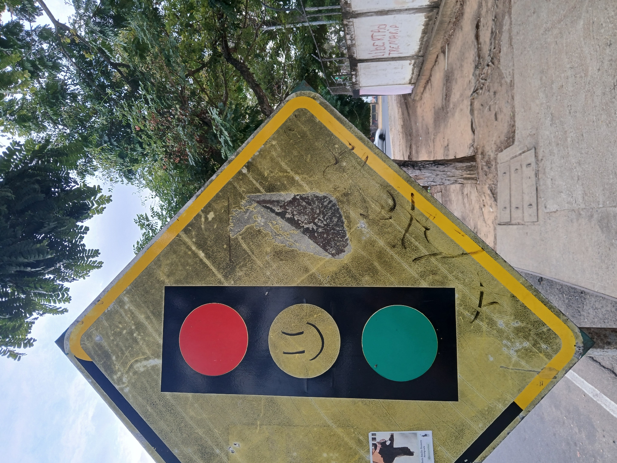 The image shows a weathered and damaged traffic signal sign in Guayana City, Venezuela. The sign is diamond-shaped with a yellow border and background. It features a black horizontal bar with three circles: a red circle on the left, a yellow circle with a smiley face in the center, and a green circle on the right. The sign shows signs of wear and tear, with peeling paint and damage in the upper portion. The sign is situated near a sidewalk, a tree, and a building with graffiti on its walls.