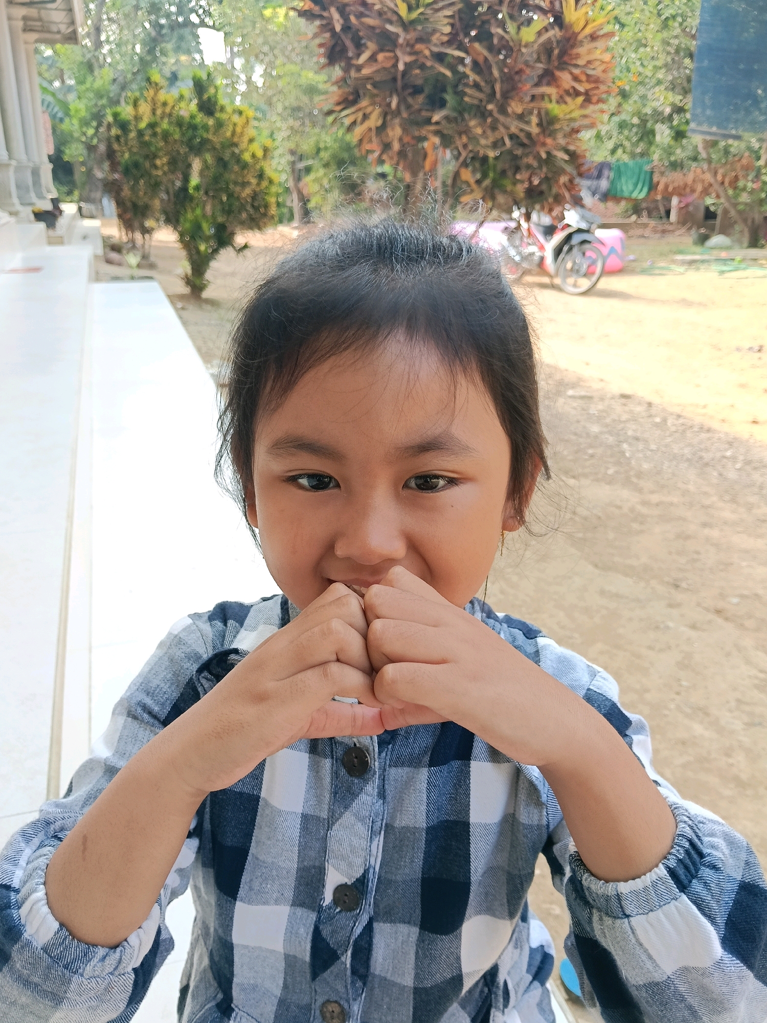 A young girl with dark hair pulled back from her face is captured in a close-up portrait, her hands clasped near her mouth. She wears a blue and white plaid shirt with puffed sleeves. The background suggests an outdoor setting with blurred greenery, trees, and what appears to be a motorcycle. The lighting is bright, indicating daylight. The girl's expression is partially obscured by her hands, but her eyes are open and directed towards the viewer.