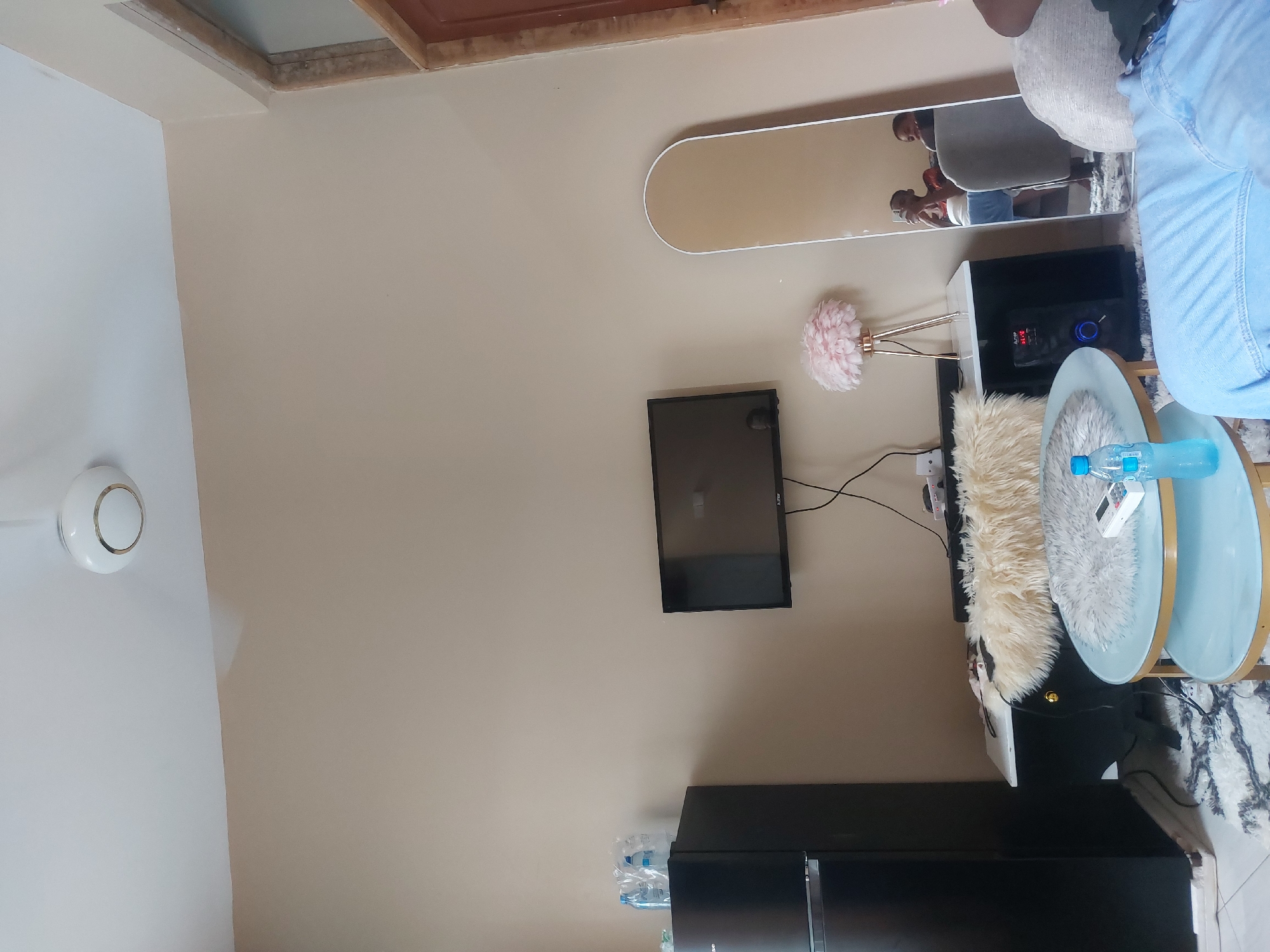 The image depicts an interior scene. A wall-mounted television is visible above a black cabinet. A decorative pink flower lamp stands near the TV. A light blue round table with a bottle of water on it is present. A mirror reflects two individuals holding phones. The room's color scheme is neutral, with beige walls and white trim. A ceiling fan is also visible. The location is unknown and cannot be determined from the image, and the reference to Dar es-Salaam, Tanzania, is not corroborated by any elements within the photograph.