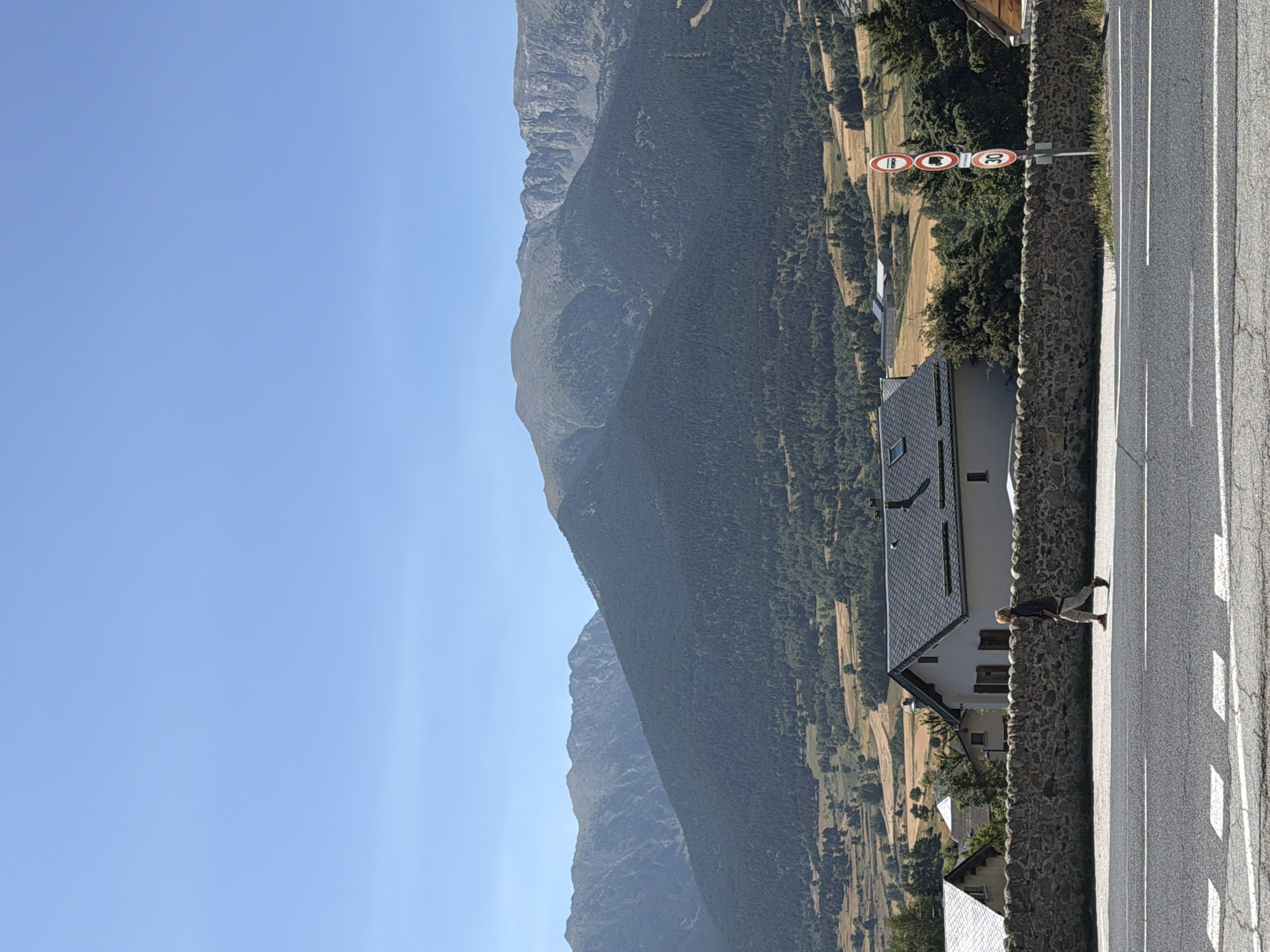 In Mont-Louis, France, a person walks along a paved road on a clear, sunny day. The road has white lane markings. Behind the road, a stone wall separates it from a residential area. In the background, there are houses with dark tiled roofs, and further back, a lush green forest covers a large hill. Beyond the hill, imposing mountains with rocky peaks rise against the blue sky. Several traffic signs, including a "30" speed limit sign, are visible on the right side of the road. The person is wearing a dark long-sleeved shirt and light-colored pants, with red shoes.