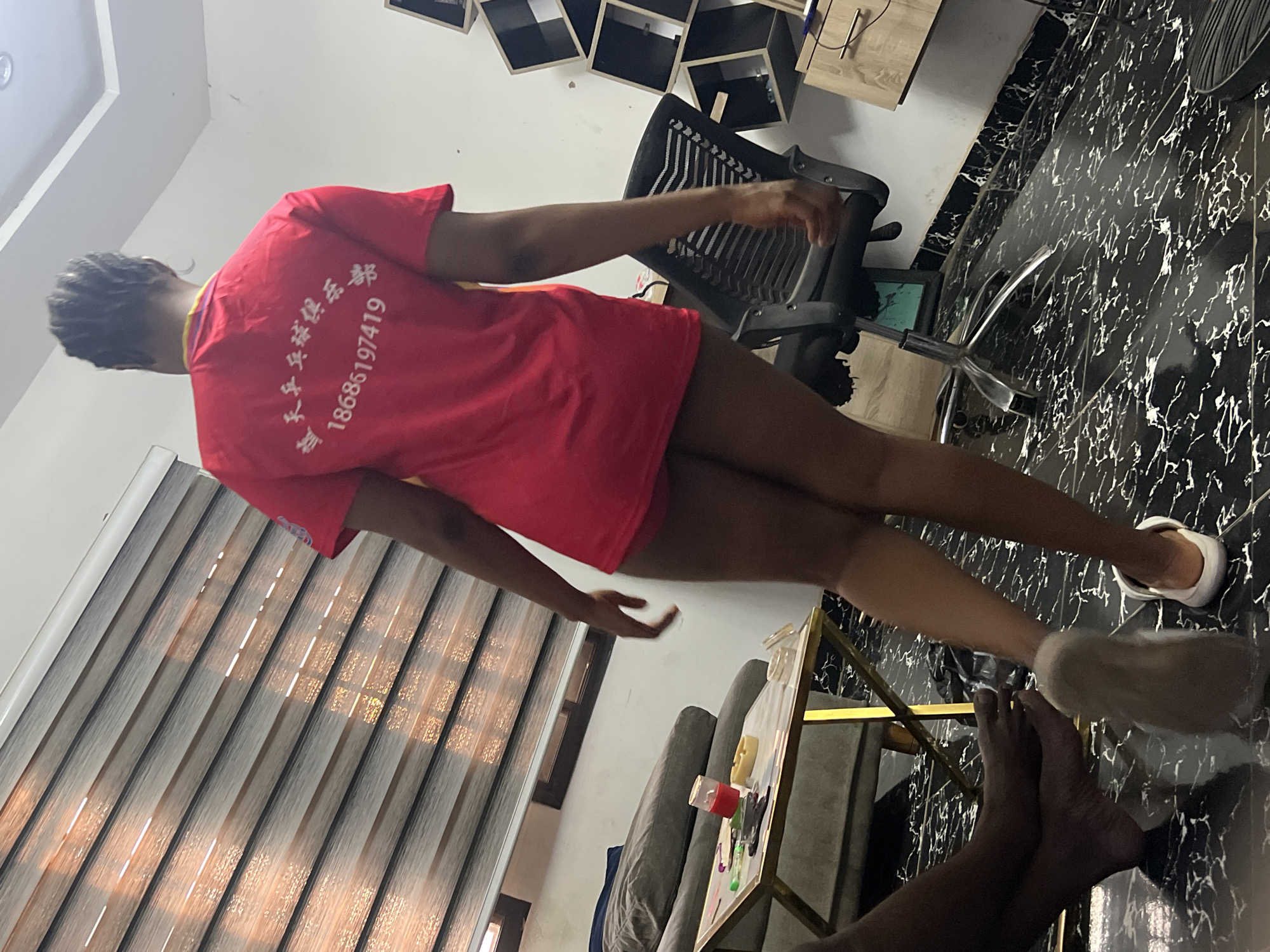 The image depicts an individual, presumably a young woman, wearing a red t-shirt with Chinese characters and the number "18686197419" printed on the back. She is standing in an interior space that appears to be a room with a patterned black and white marble floor, blinds covering a window, and a desk chair. There are items such as a small desk or table with art supplies and a grey sofa visible, as well as what appear to be decorative black boxes mounted on a wall. Another person’s feet are visible in the foreground of the photo. The room is located within the city of Abuja, Nigeria.