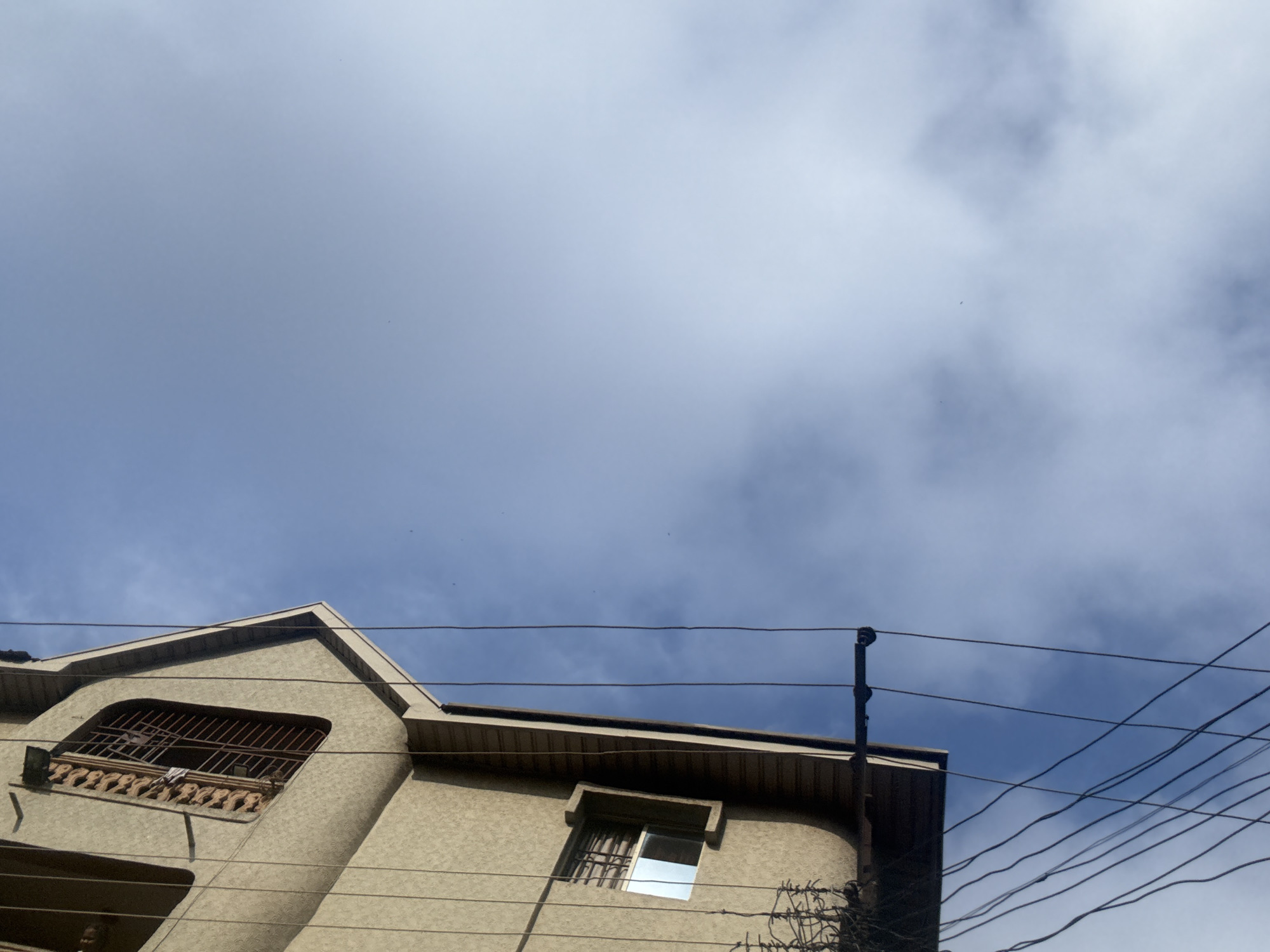 This image captures a close-up, low-angle view of a building in Onitsha, Nigeria, against a backdrop of a blue sky with wispy clouds. The building, with its textured, light brown facade, occupies the left side of the frame. A prominent window with a decorative, metal grate is visible on the upper section of the building. Below it, a smaller, rectangular window shows a reflection on its glass. Several dark, overhead electrical or telecommunication wires crisscross the scene, adding a sense of urban density. The perspective emphasizes the upward expanse of the sky, with a portion of the building's roofline visible at the top left. The overall impression is one of everyday urban infrastructure and natural elements juxtaposed.