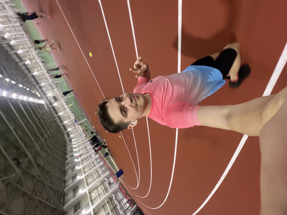 An indoor selfie captures a young man standing on a reddish-brown running track with white lane lines. He is positioned in the foreground, holding the camera with his extended left arm, capturing himself from a slightly low angle. He has short, dark hair and is wearing a pink t-shirt that transitions into light blue shorts, with black compression shorts visible underneath. White wireless earbuds are in his ears. He looks directly at the camera with a relaxed expression, a slight smile on his face, and his right hand is raised slightly, fingers in a loose gesture.
In the background, further down the track, several other individuals, presumably athletes, are scattered. Some appear to be standing or stretching, and others are near hurdles that are set up on the track, suggesting a training session or athletic event. Small yellow and pink disc markers are also visible on the track surface.
The setting is a large indoor athletic facility or gymnasium, indicated by the architecture in the background on the left side of the frame. The walls feature multiple levels of windows or open structures, and the space is illuminated by artificial lighting, characteristic of an indoor sports complex. The overall atmosphere appears active and energetic, consistent with a sports training environment.