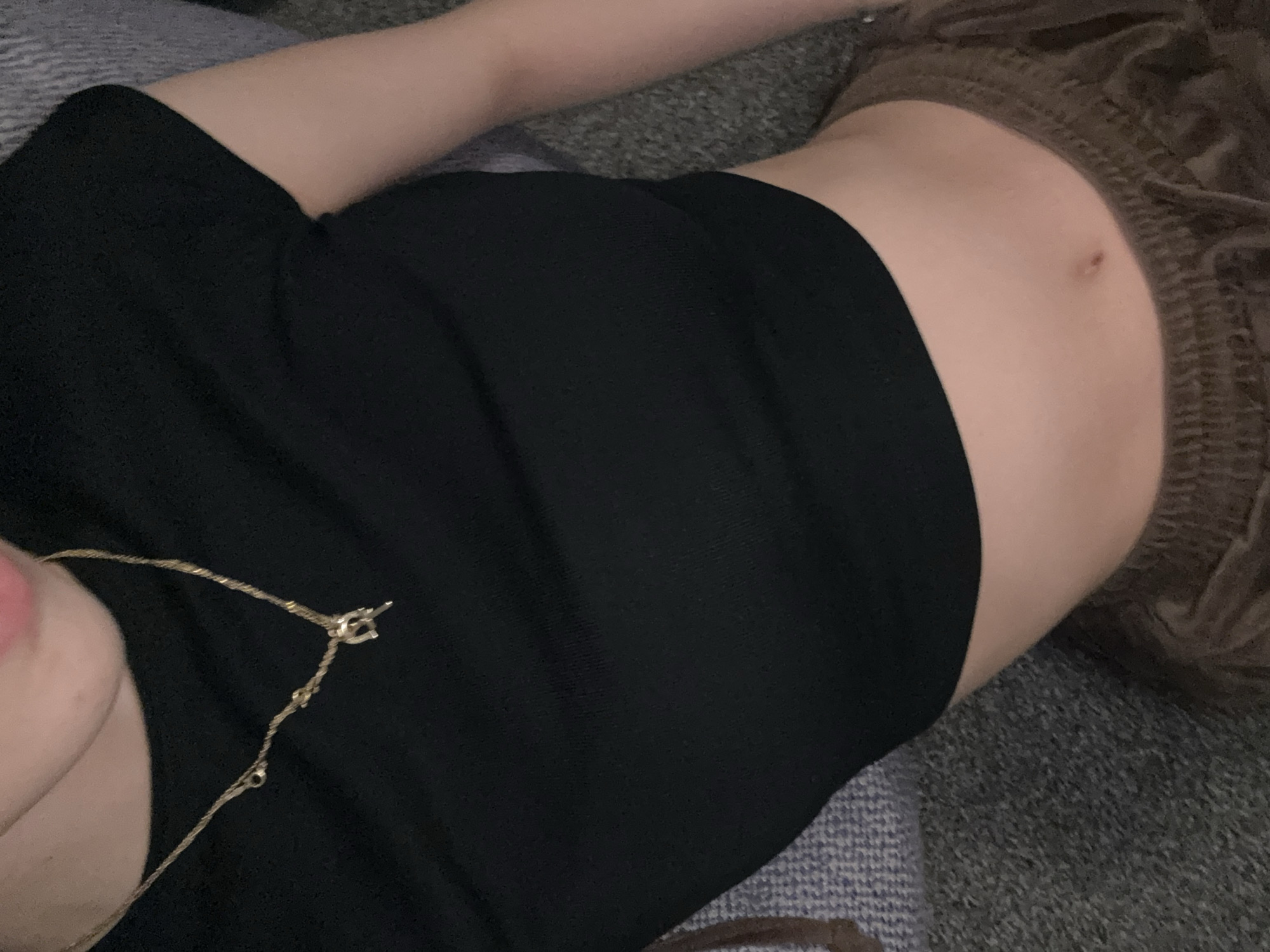 The image shows a person lying down, wearing a black top. Part of their abdomen is visible. A gold necklace is around their neck. The person is lying on a gray surface, possibly a couch or bed. A brown, textured fabric is visible on the right side of the image. The location is not determinable from the provided image.