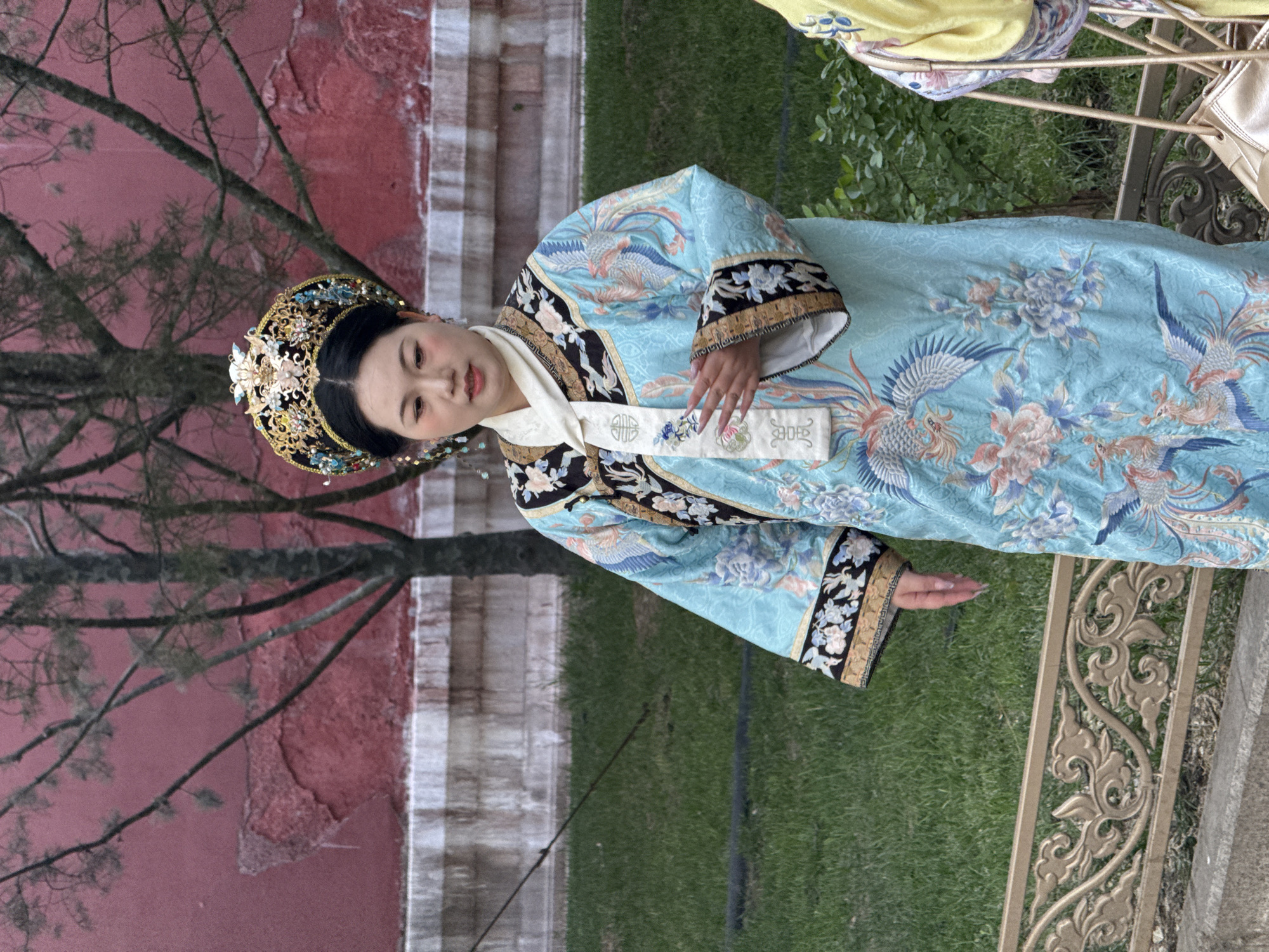 A woman in traditional Qing Dynasty clothing stands in what appears to be a courtyard. The woman wears an ornate headdress, a light blue robe adorned with phoenix and floral patterns, and has long fingernails. Behind her is a red wall with peeling paint, a bare tree, and a set of stone steps. The ground is covered in green grass and a decorative metal railing is visible on the right side of the image. The image was taken in the Dongcheng District, China.