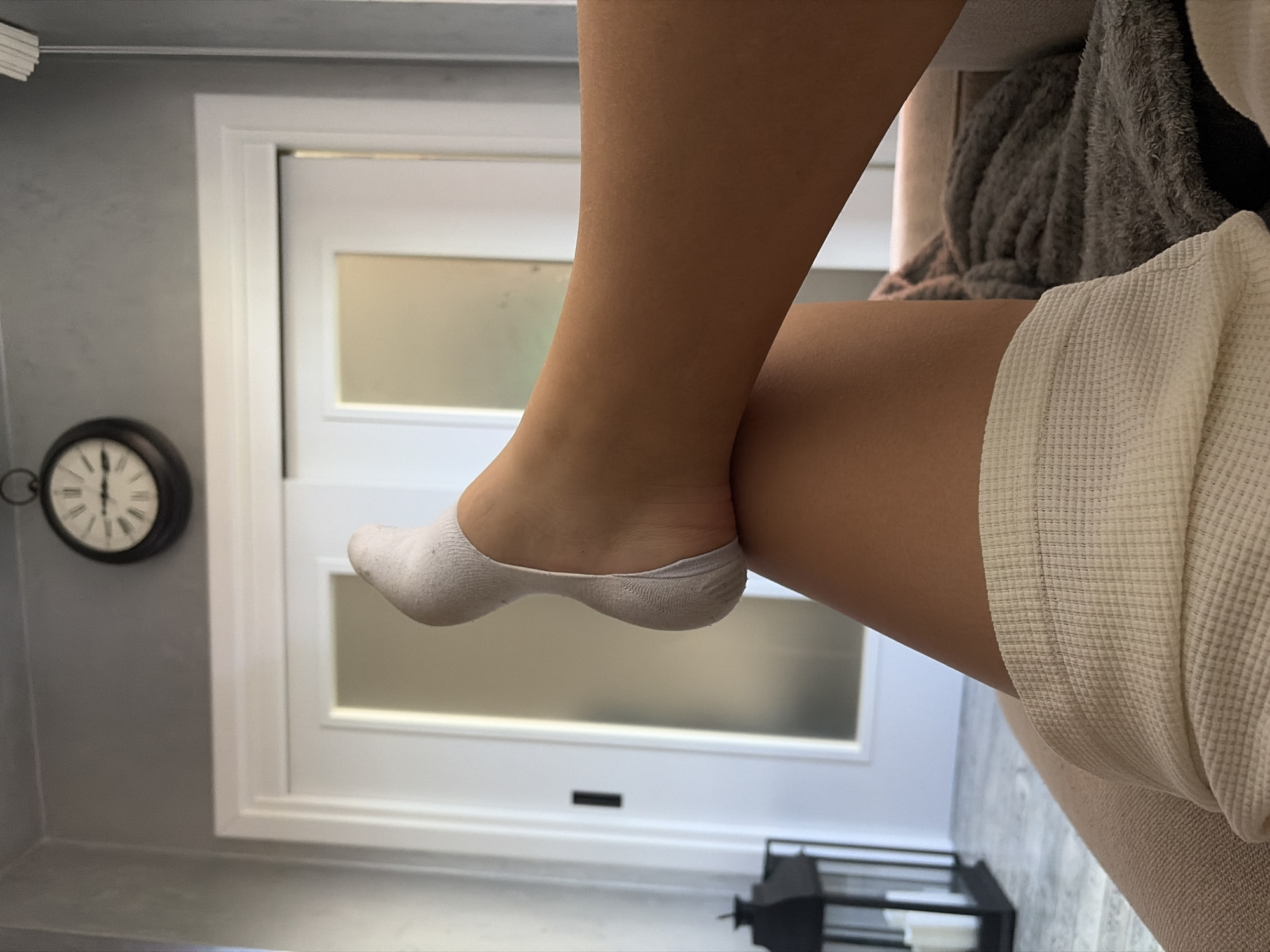 A close-up, angled shot focuses on a person's lower leg and foot wearing a white no-show sock. The leg is bent, with the foot positioned in the foreground. The person is wearing a textured, light-colored garment, possibly a robe or blanket, that obscures part of the background.
Behind the leg and foot is a white door with a horizontal rectangular panel in the center. To the left of the door, partially visible, is a black wall-mounted clock with Roman numerals. In the lower right corner, a decorative black metal object is visible.
The image does not contain enough information to determine the location in Shumen, Bulgaria.