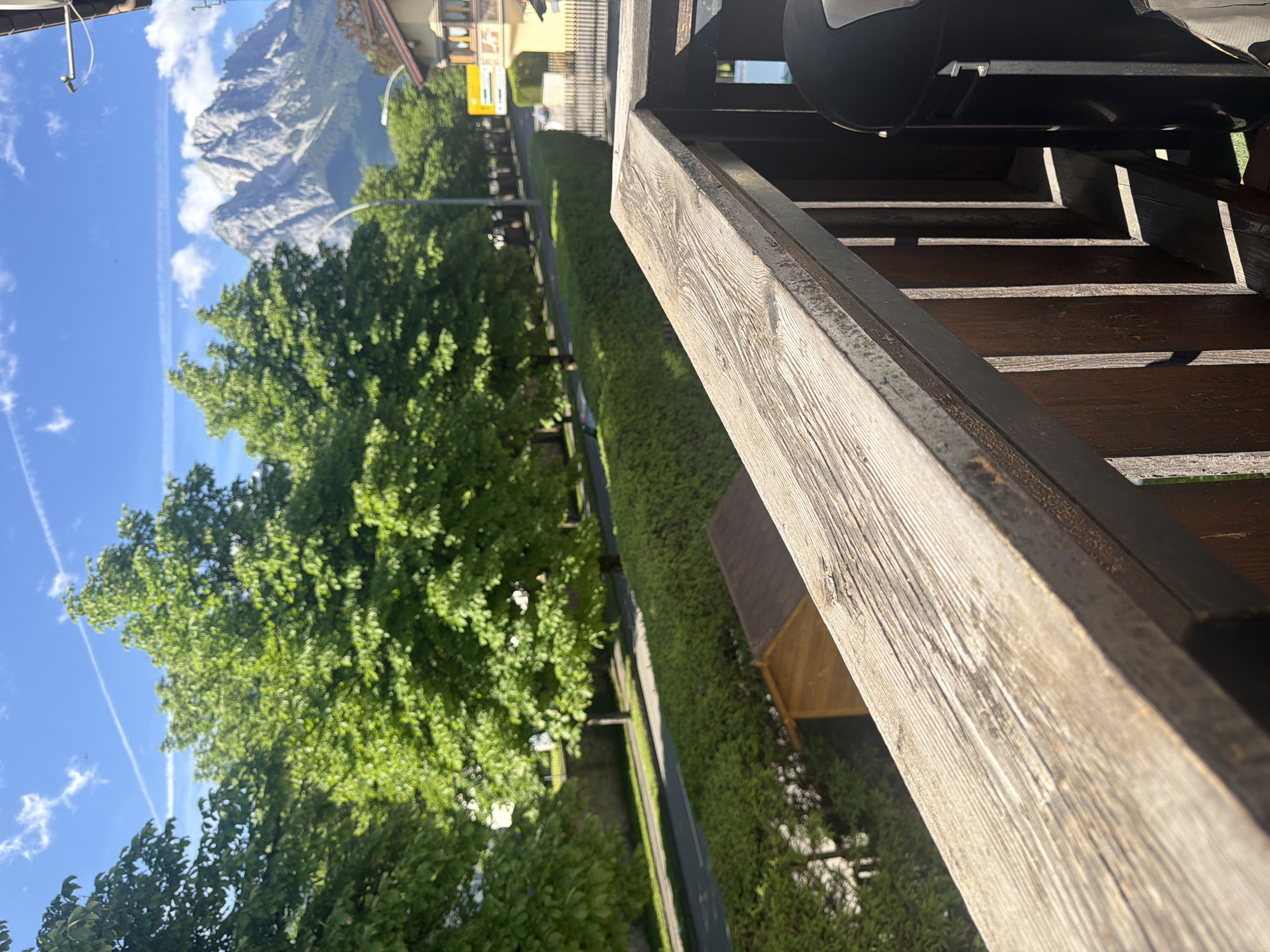 The image shows a view from a wooden balcony, overlooking a street lined with trees and a distant mountain range. The balcony railing in the foreground is made of weathered wood, with dark brown wooden posts supporting it. Beyond the railing, a neat green hedge runs along the side of a road. A small wooden shed is visible behind the hedge. The road itself is empty of traffic. In the background, large, lush green trees form a canopy, with a bright blue sky dotted with white clouds and airplane contrails above. The majestic, rocky peaks of mountains dominate the far distance, under the bright sunlight. A yellow sign is visible on the right side of the image, and part of a building with a balcony is also seen.