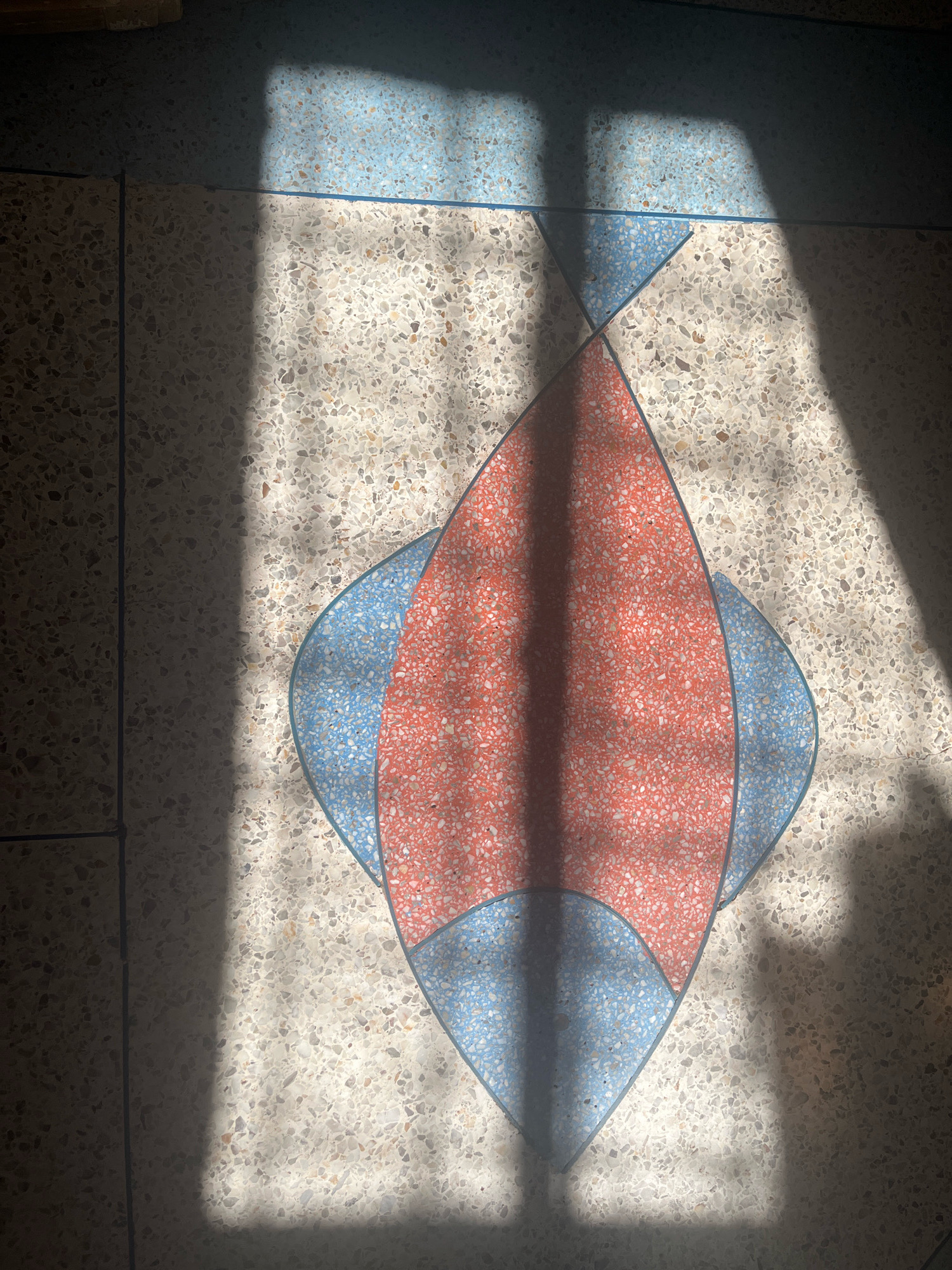 The image shows a close-up, overhead view of a patterned floor with terrazzo tiles. The floor features a central diamond-shaped design with sections of reddish-orange and light blue terrazzo, surrounded by cream-colored terrazzo flecked with various neutral tones. The pattern is outlined with thin dark lines. Sunlight casts distinct shadows across the floor, creating geometric shapes and obscuring parts of the pattern, suggesting an indoor environment with sunlight streaming through a window or similar opening. The location is identified as Azilal, Morocco.