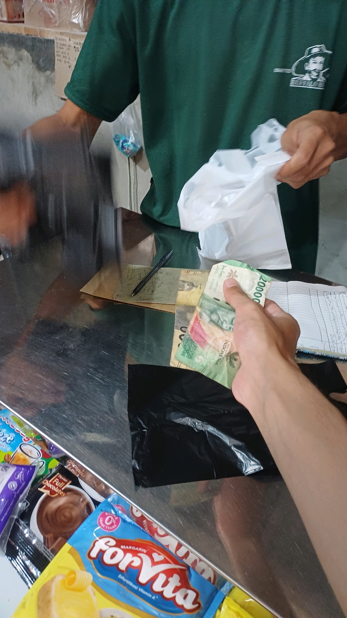 In a retail setting, possibly a small store or market stall in Kranggan, Indonesia, a transaction is occurring. A person, whose right arm and hand are visible, is holding several Indonesian Rupiah banknotes, including a 20,000 Rupiah note. The hand appears to be in the process of paying or receiving money. In the foreground, there are several packaged food items, including a blue and yellow package labeled "for Vita Margarin" and other snack items. A black plastic bag is also present on the counter. Behind the counter, a person wearing a dark green t-shirt is holding a white plastic bag. The counter surface is reflective metal. The background shows shelves and a wall with some items on them. The overall scene suggests a typical point-of-sale interaction.