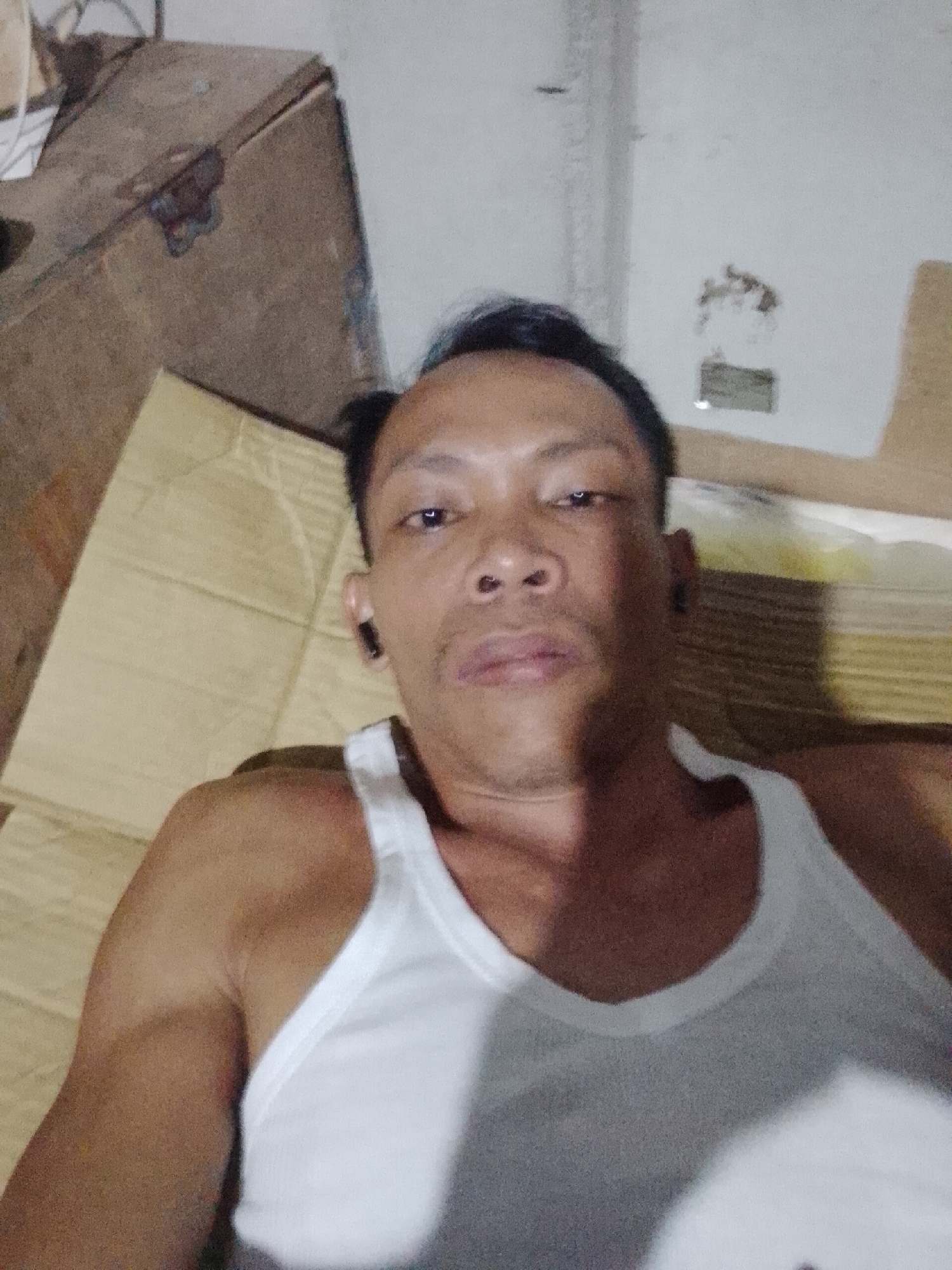 A selfie of a man wearing a white tank top in an indoor environment. He is looking at the camera. To the left, there is a wooden chest or box. Behind him and to the right is a wall with a light fixture. The location is Bukit Ketok, Indonesia. There is corrugated cardboard on the floor. The man has short, dark hair and appears to have earbuds in his ears.